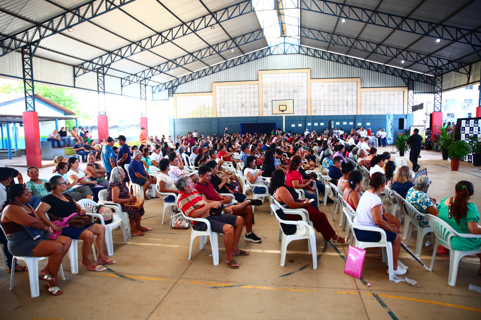 Foto colorida mostra uma grande quadra poliesportiva coberta, completamente ocupada por um público sentado em cadeiras de plástico brancas. A maioria dos presentes é de adultos. Foto colorida mostra uma grande quadra poliesportiva coberta, completamente ocupada por um público sentado em cadeiras de plástico brancas. A maioria dos presentes é de adultos.