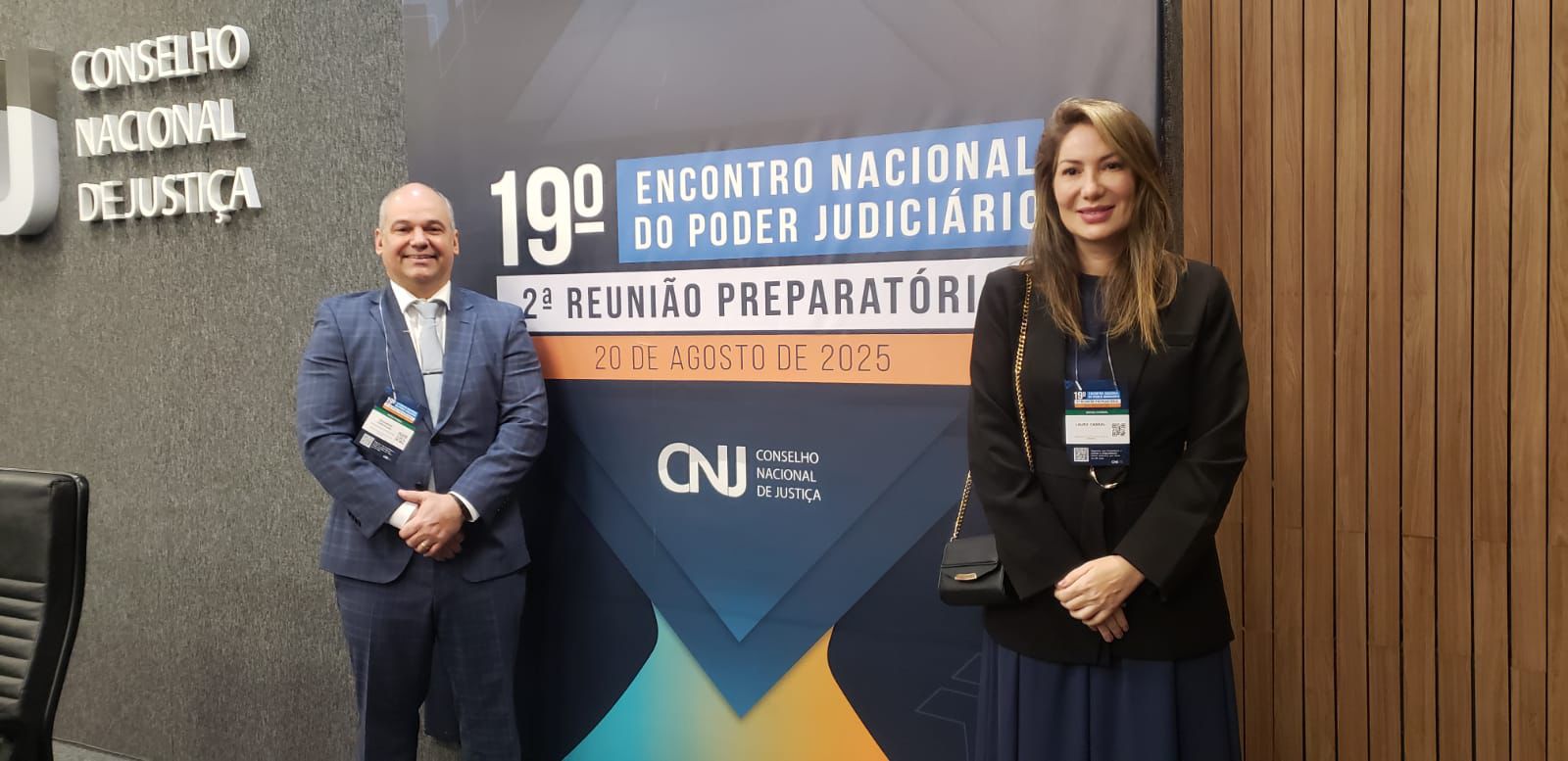  Imagem colorida mostra duas pessoas, um homem e uma mulher, em pé em frente a um banner. O homem, careca e de terno azul-marinho, usa um crachá no peito. A mulher, com cabelo castanho solto, usa um blazer preto e uma saia longa azul. O banner tem um logo que diz "Conselho Nacional de Justiça" e o título "19º Encontro Nacional do Poder Judiciário, 2ª Reunião Preparatória, 20 de agosto de 2025".