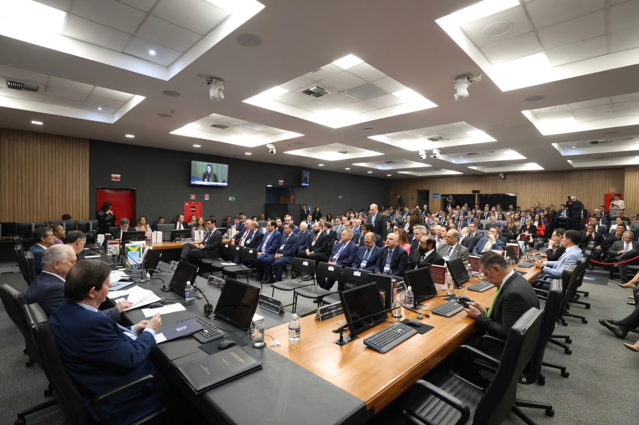 Imagem colorida mostra uma sala de conferências grande, com muitas pessoas sentadas em fileiras de cadeiras e outras em uma mesa comprida. A maioria das pessoas está vestindo ternos. Há computadores, notebooks e garrafas de água sobre a mesa principal. No centro da sala, um telão mostra uma mulher em um vídeo. Imagem colorida mostra uma sala de conferências grande, com muitas pessoas sentadas em fileiras de cadeiras e outras em uma mesa comprida. A maioria das pessoas está vestindo ternos. Há computadores, notebooks e garrafas de água sobre a mesa principal. No centro da sala, um telão mostra uma mulher em um vídeo.