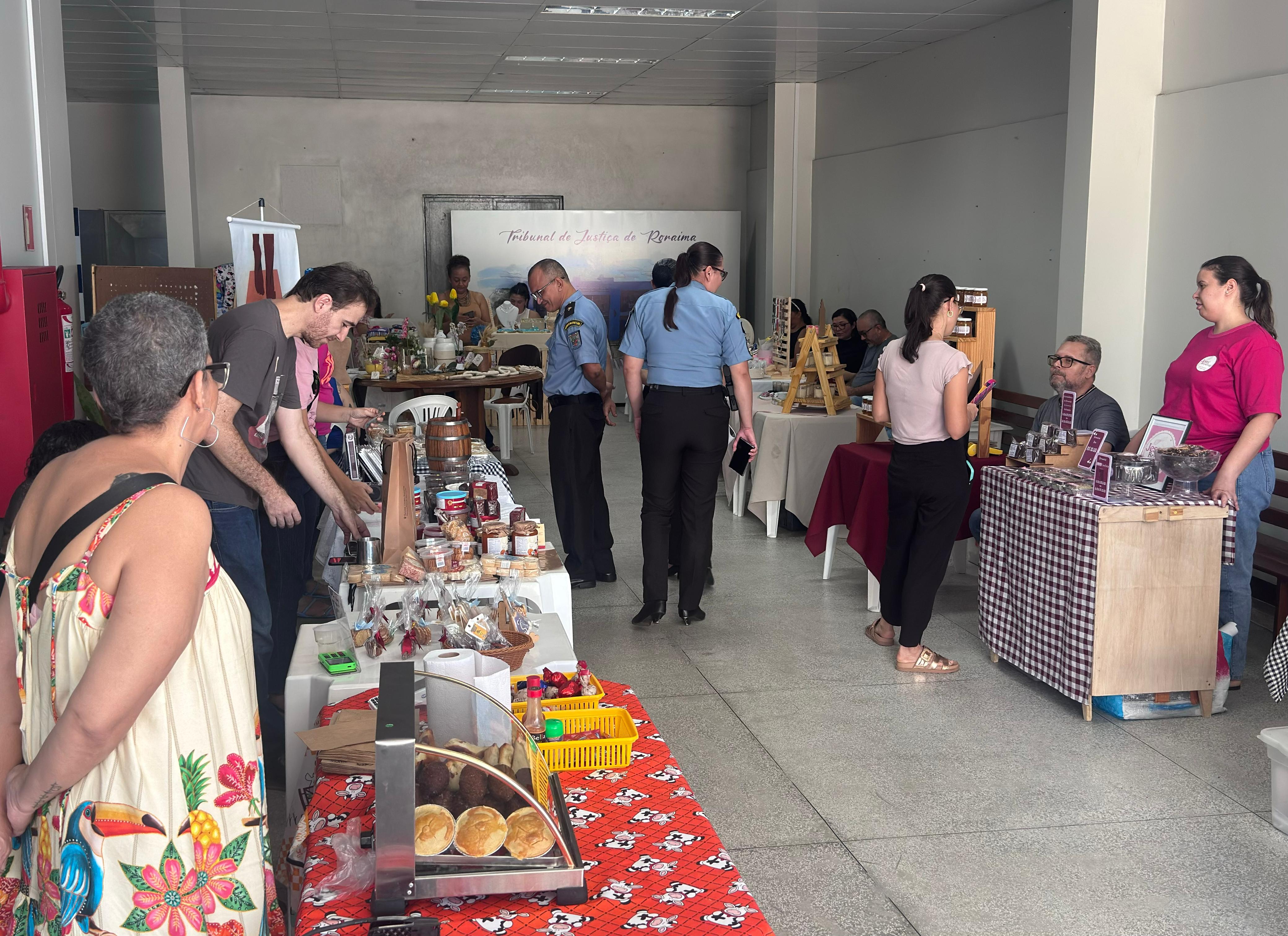 Imagem colorida mostra um salão de eventos, com pessoas circulando entre várias barracas de artesanato e comida. No primeiro plano, uma barraca exibe doces e pães sobre uma toalha vermelha com estampa de galinhas. Ao fundo, outras barracas exibem produtos diversos e há pessoas conversando e comprando. Duas pessoas vestidas com uniformes azuis e pretos circulam entre as barracas. Imagem colorida mostra um salão de eventos, com pessoas circulando entre várias barracas de artesanato e comida. No primeiro plano, uma barraca exibe doces e pães sobre uma toalha vermelha com estampa de galinhas. Ao fundo, outras barracas exibem produtos diversos e há pessoas conversando e comprando. Duas pessoas vestidas com uniformes azuis e pretos circulam entre as barracas.
