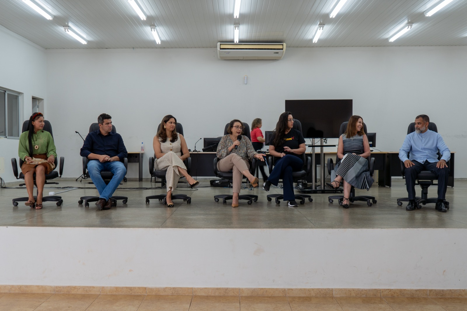 Item 3: fotografia tirada de frente, mostra seis pessoas adultas sentadas em cadeiras pretas de escritório sobre um palco. Elas estão em uma fileira, em frente a um grande painel de televisão. Todas as pessoas estão com roupas sociais ou casuais.