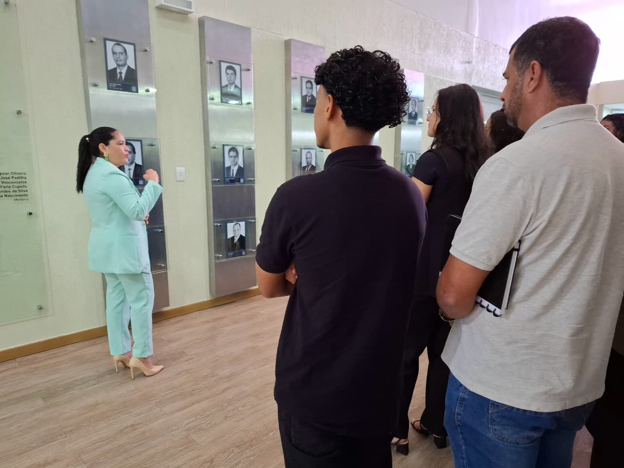 Imagem colorida mostra uma mulher de costas para a câmera, usando um terno azul-claro, sapatos de salto e fones de ouvido. Ela está em pé, de lado, gesticulando com a mão direita e olhando para um grupo de pessoas que a observa. Eles estão em uma sala com paredes claras, onde há várias placas de vidro com retratos dos ex-presidentes do TJRR.