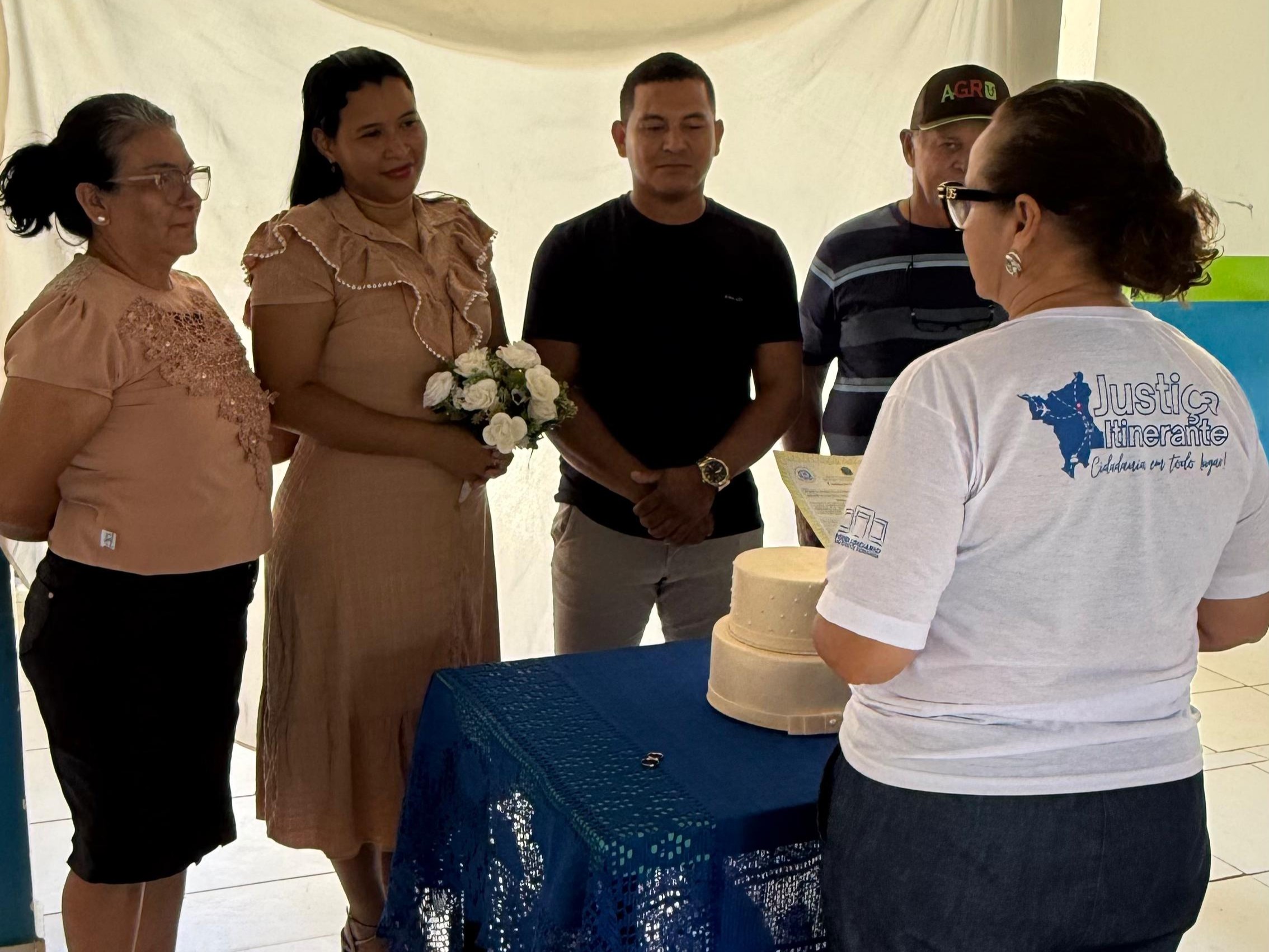 Foto colorida mostra um casamento civil. Uma mulher de vestido rosa claro, segurando um buquê de flores brancas, e um homem de camiseta preta e calça cinza estão no centro, de frente para a juíza Graciete Sotto Mayor, titular da Vara da Justiça Itinerante que está vestindo uma camiseta branca com o logo "Justiça Itinerante". Essa mulher está conduzindo a cerimônia. À esquerda do casal, uma mulher de blusa rosa e saia preta observa a cena. À direita do casal, outro homem com boné e camiseta listrada também acompanha. Uma pequena mesa coberta com uma toalha de renda azul escura está entre a juíza e o casal, e sobre ela há um pequeno bolo de casamento e as alianças.