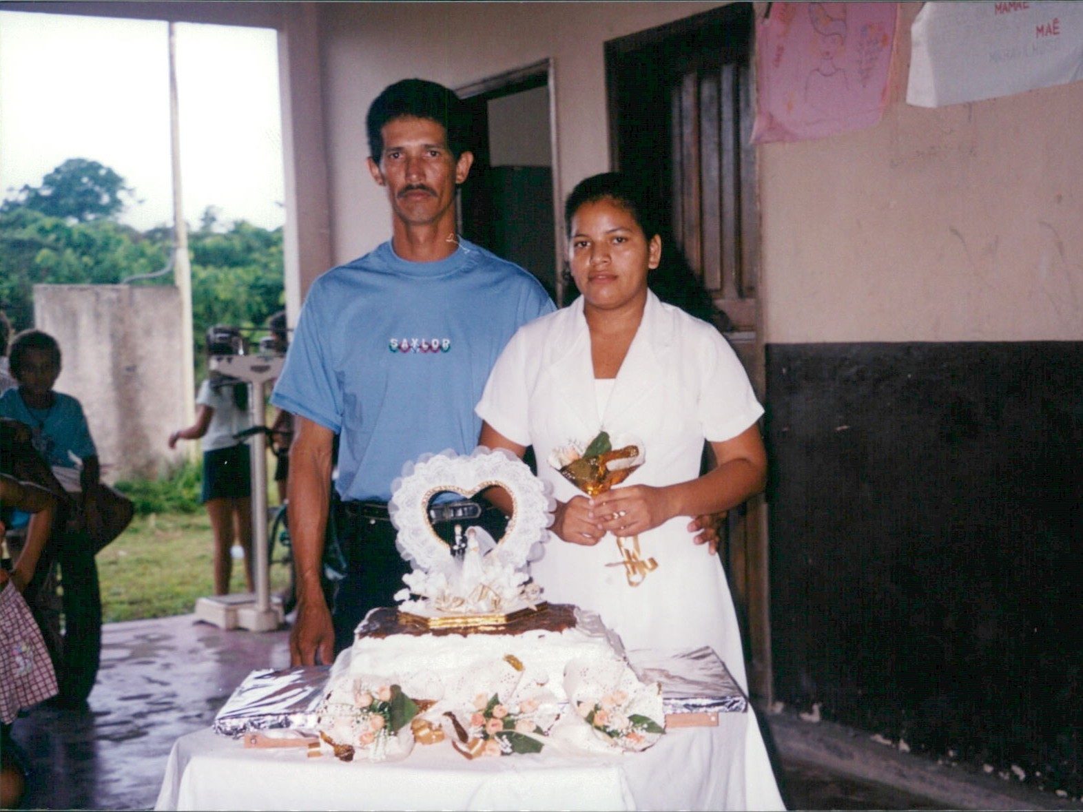 Um homem e uma mulher estão em pé, lado a lado, atrás de uma mesa decorada com um bolo de casamento simples e enfeitado com flores, laços e um topo com bonequinhos de noivos dentro de um coração. O homem, de pele clara, bigode e cabelos curtos, veste uma camiseta azul e calça jeans. A mulher, de pele morena e cabelos presos, veste um vestido branco e segura um pequeno buquê dourado. Eles olham para a câmera com expressões serenas.