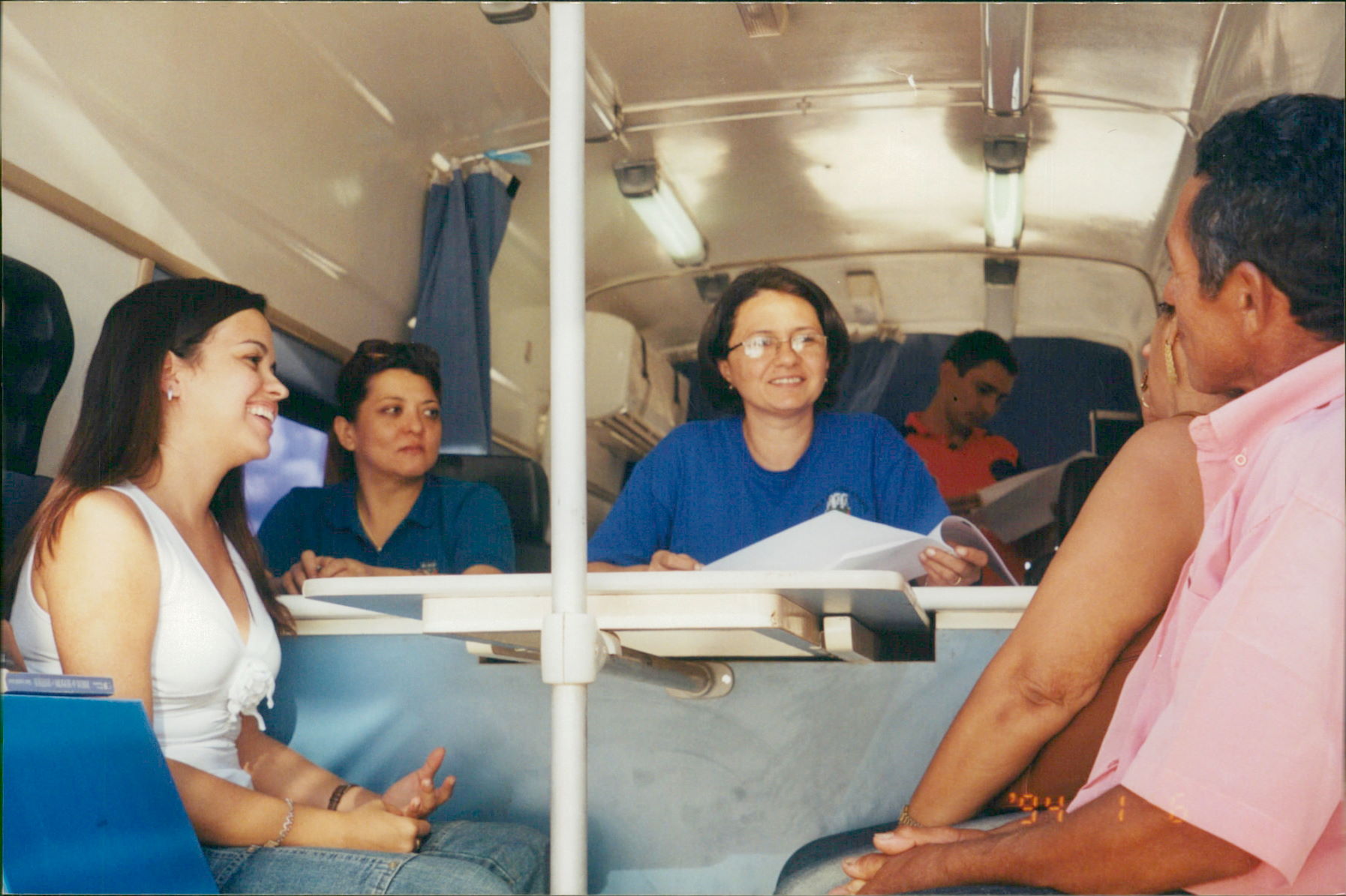  Imagem colorida mostra três mulheres e dois homens sentados dentro de um veículo. Uma mulher de camiseta azul no centro olha para a frente. Uma mulher de camiseta branca e um homem de camiseta rosa se olham e sorriem. Outra mulher de camiseta azul escura está sentada ao fundo. Um homem com camiseta laranja está sentado atrás da mulher de azul no centro