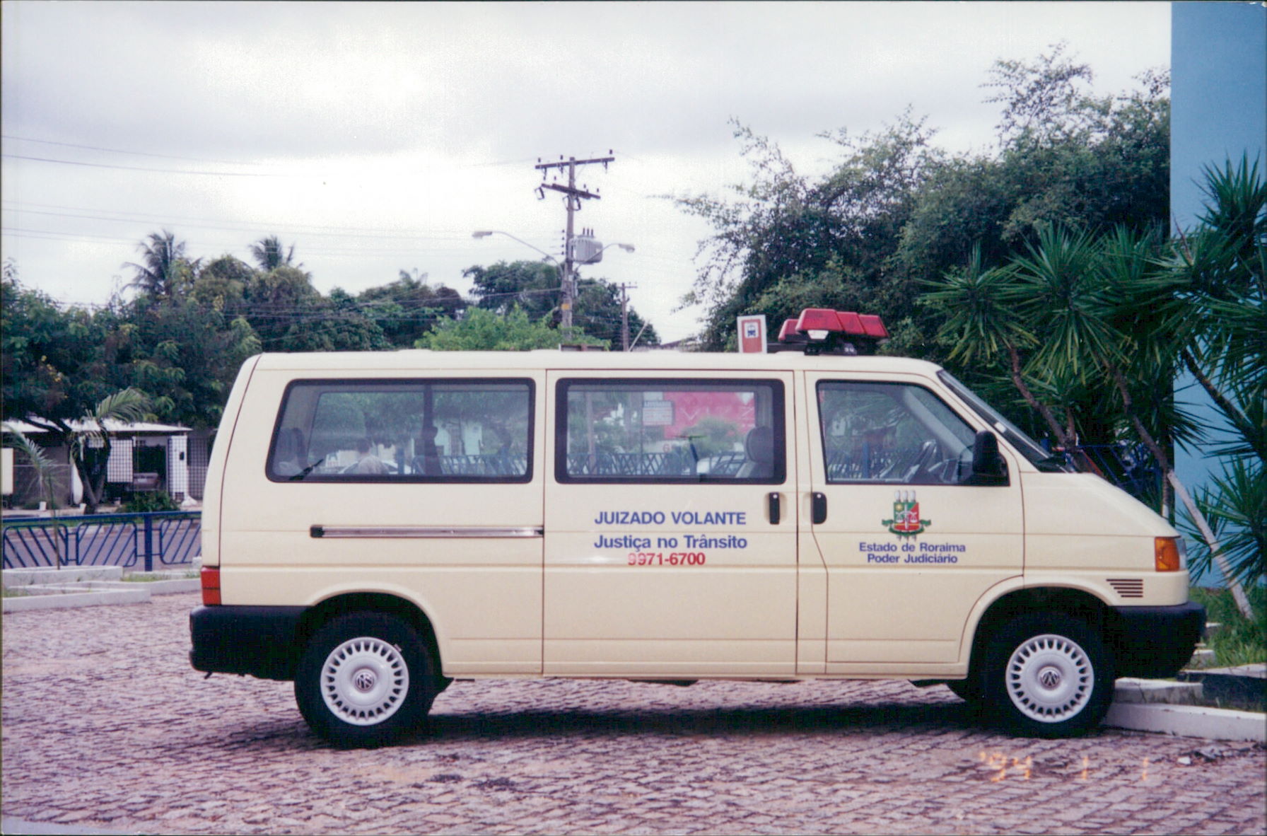 Imagem colorida mostra uma van bege com o letreiro "Juizado Volante Justiça no Trânsito" está estacionada em uma área com calçamento e vegetação ao fundo. O brasão do "Estado de Roraima Poder Judiciário" está estampado na lateral do veículo.