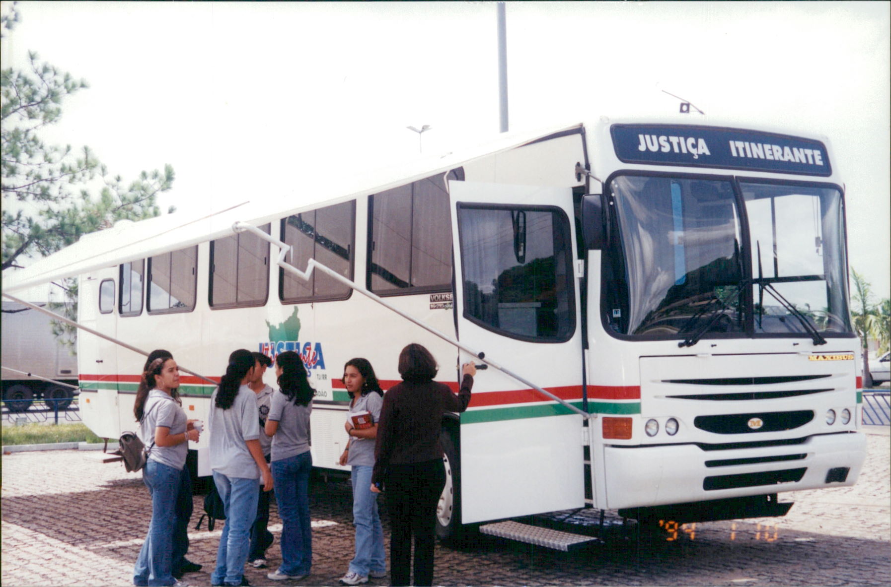 Imagem colorida mostra um ônibus branco com faixas vermelhas e verdes, com a inscrição "Justiça Itinerante" na parte superior, que está estacionado em uma área pavimentada. A porta da frente está aberta e um grupo de quatro jovens e uma mulher estão próximos a ela.