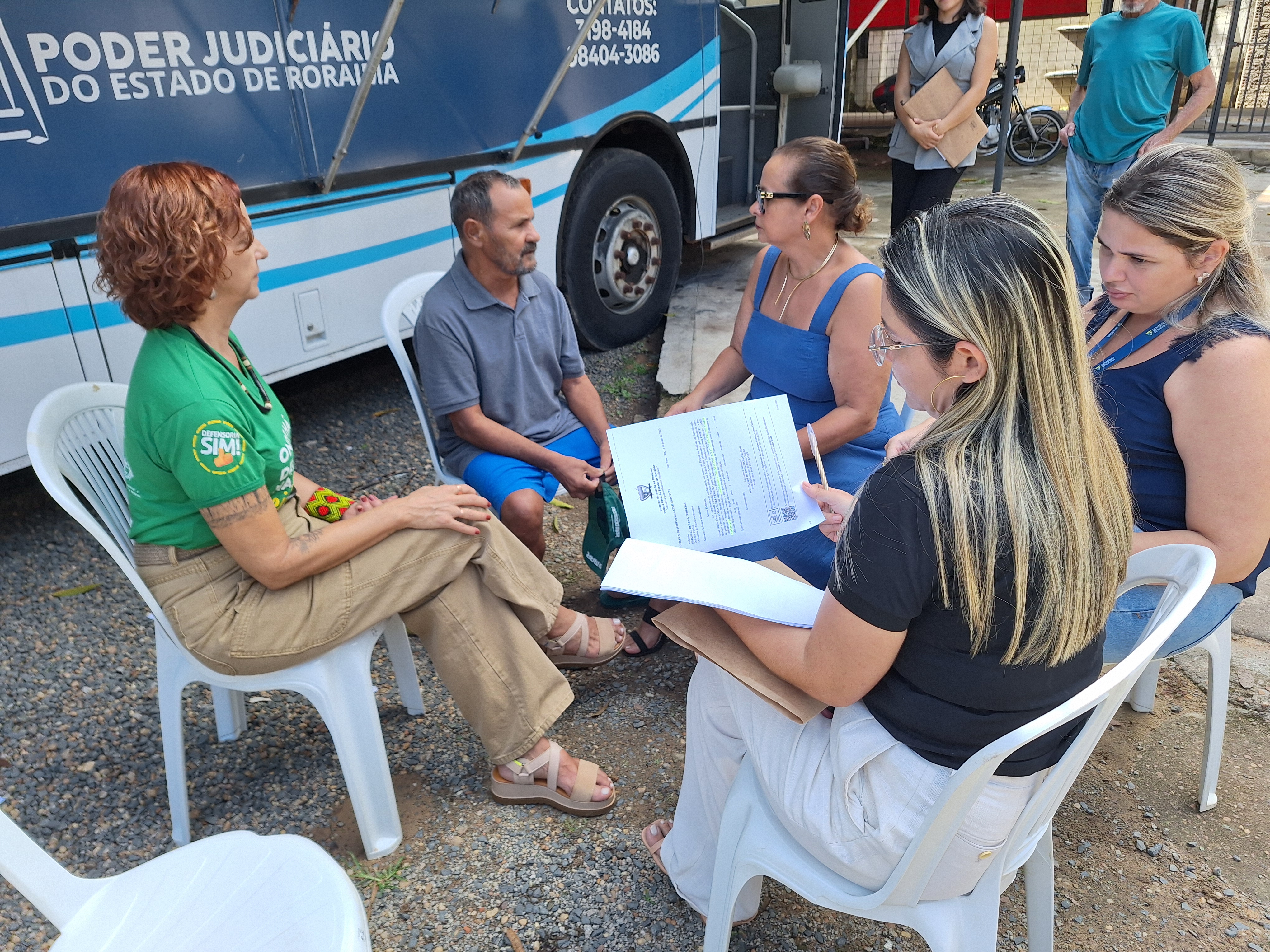 Em um ambiente externo, seis pessoas participam de um atendimento jurídico sob a estrutura da Justiça Itinerante. Elas estão sentadas em cadeiras plásticas brancas, formando uma roda de conversa.