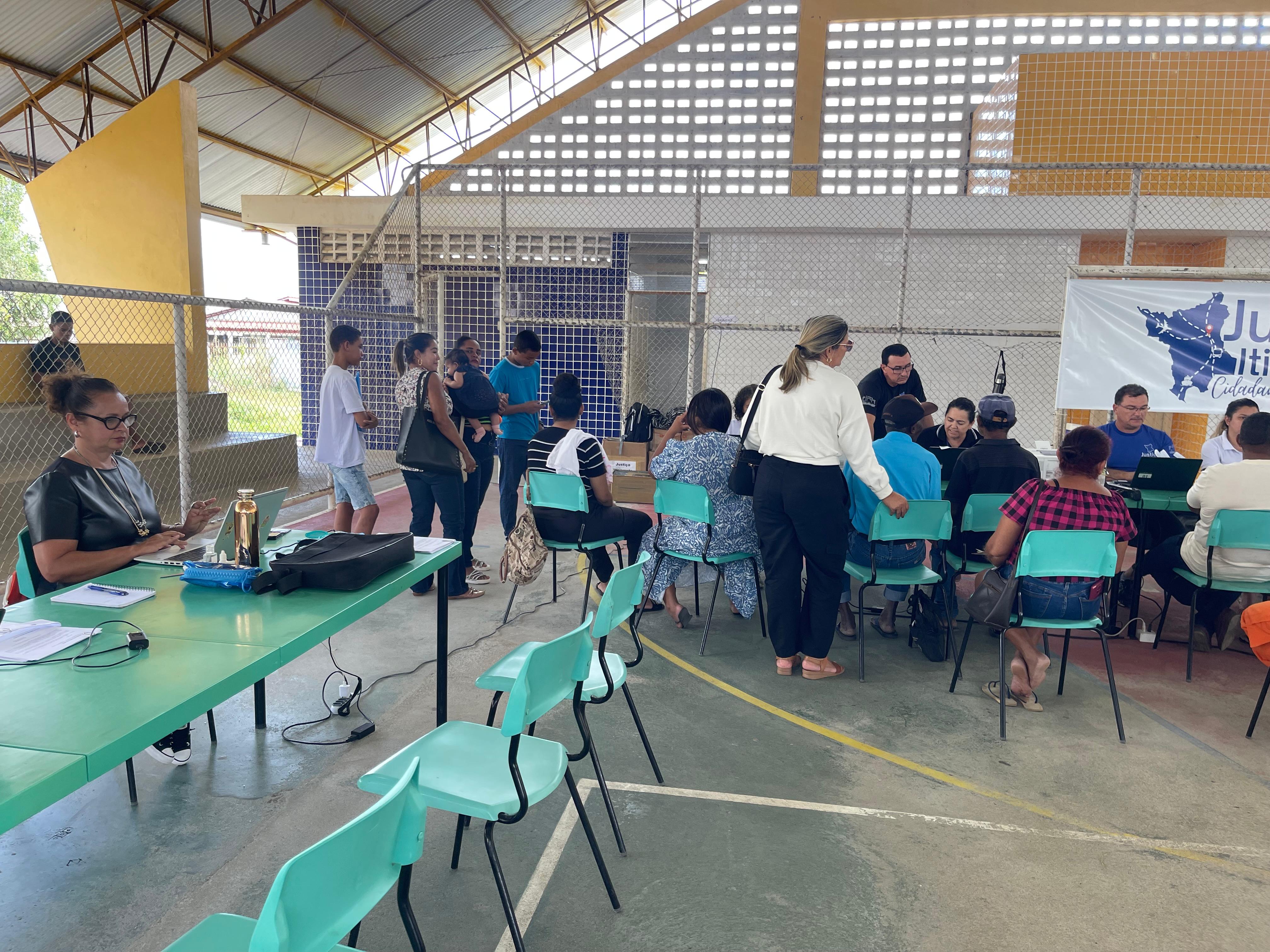 A foto mostra um mutirão da Justiça Itinerante acontecendo dentro de uma quadra coberta. À esquerda, uma mulher de óculos trabalha sozinha em uma mesa verde com caderno, celular, garrafa térmica e bolsas. À direita, uma fileira de pessoas está sentada diante de atendentes — alguns com camisetas azuis da Justiça Itinerante. Ao fundo, há um banner branco com o mapa de Roraima em azul e os dizeres “Justiça Itinerante – Cidadania em Movimento”. Algumas pessoas estão de pé, aguardando atendimento, incluindo uma mulher com bebê no colo. A estrutura da quadra é cercada por grade e parcialmente coberta com telhado metálico.