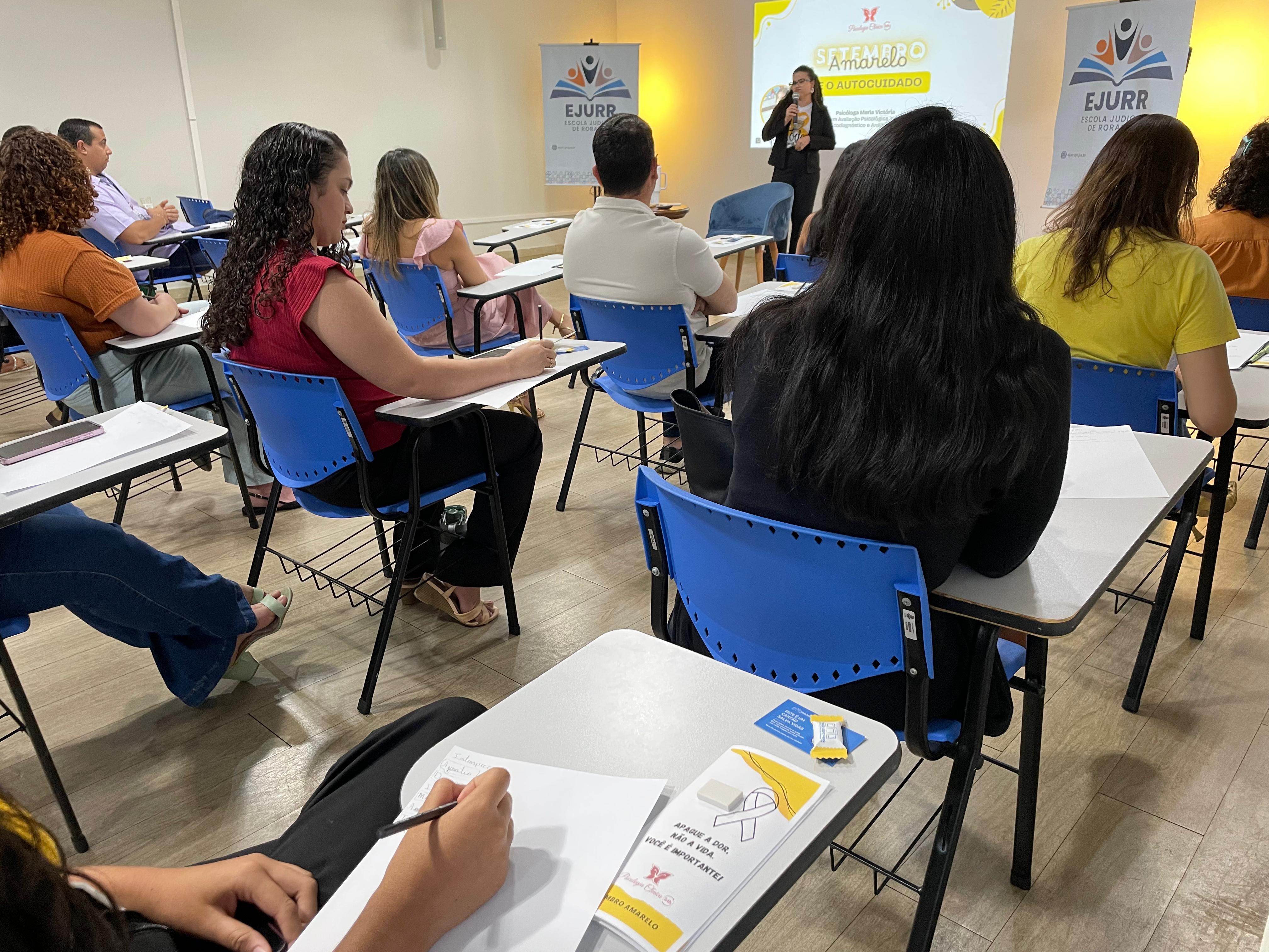 Foto colorida mostra uma sala com diversas pessoas sentadas em cadeiras escolares azuis, escrevendo em folhas de papel. À frente, uma mulher em pé realiza uma palestra com o tema "Setembro Amarelo e o Autocuidado", conforme apresentado no telão.