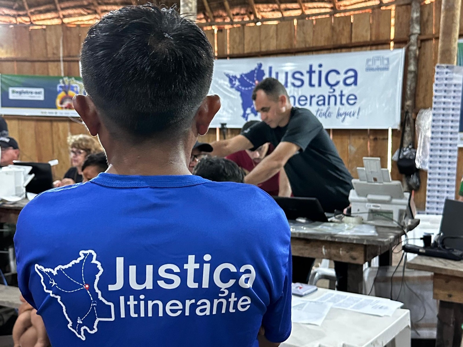  A imagem colorida mostra um ambiente interno de um local de atendimento, possivelmente uma sala rústica de madeira. Um homem de costas, vestindo uma camiseta azul com a estampa “Justiça Itinerante”, está em primeiro plano. Ao fundo, outras pessoas estão sentadas e um homem de camiseta preta está inclinado sobre uma mesa com um computador, uma impressora e papéis. Há um banner ao fundo com a inscrição “Justiça Itinerante” e o mapa de Roraima.