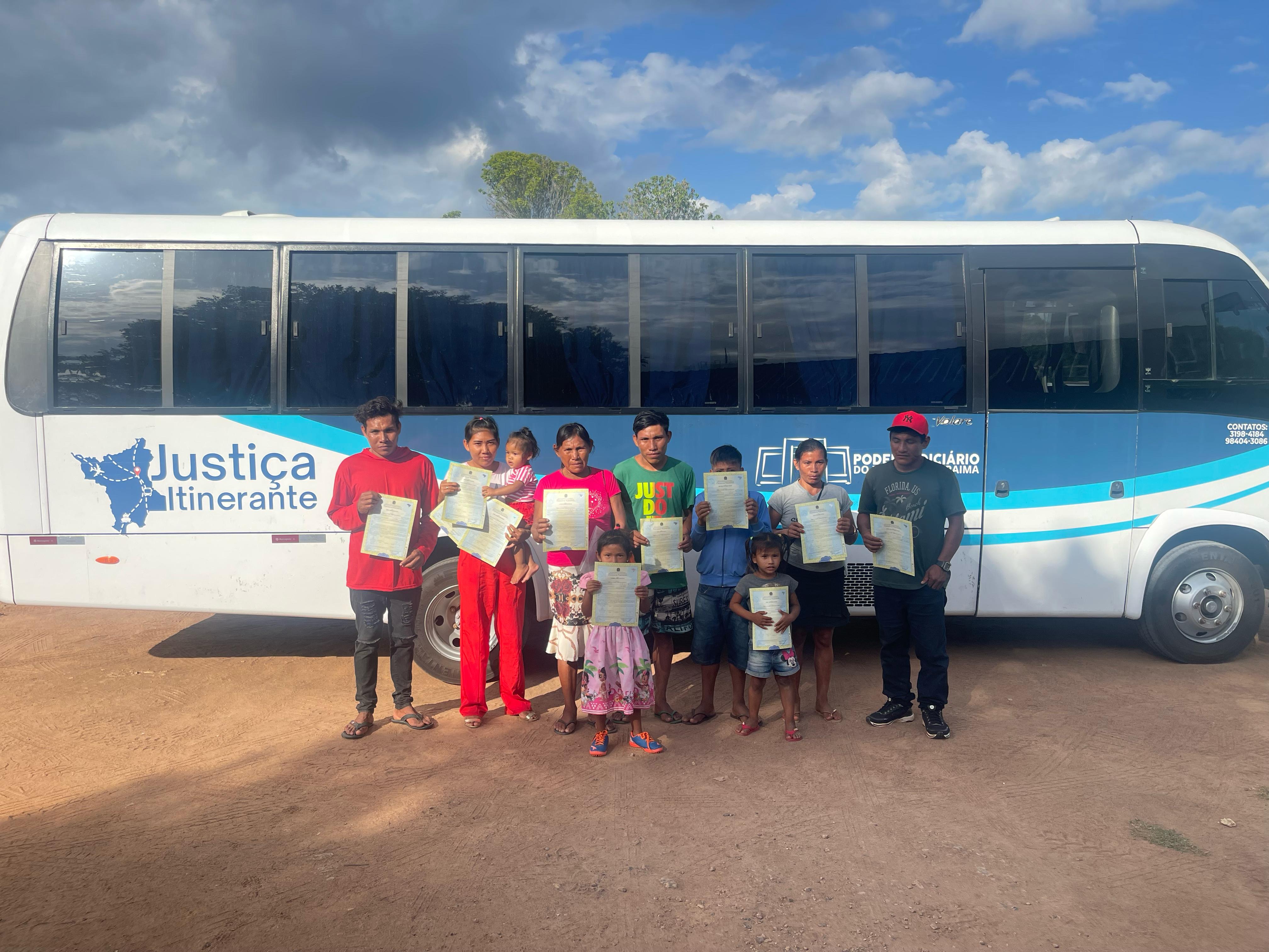 Imagem colorida mostra um grupo de adultos e crianças segurando documentos, em frente a um ônibus branco e azul com a inscrição "Justiça Itinerante". Todos estão em uma área de terra batida sob um céu parcialmente nublado.