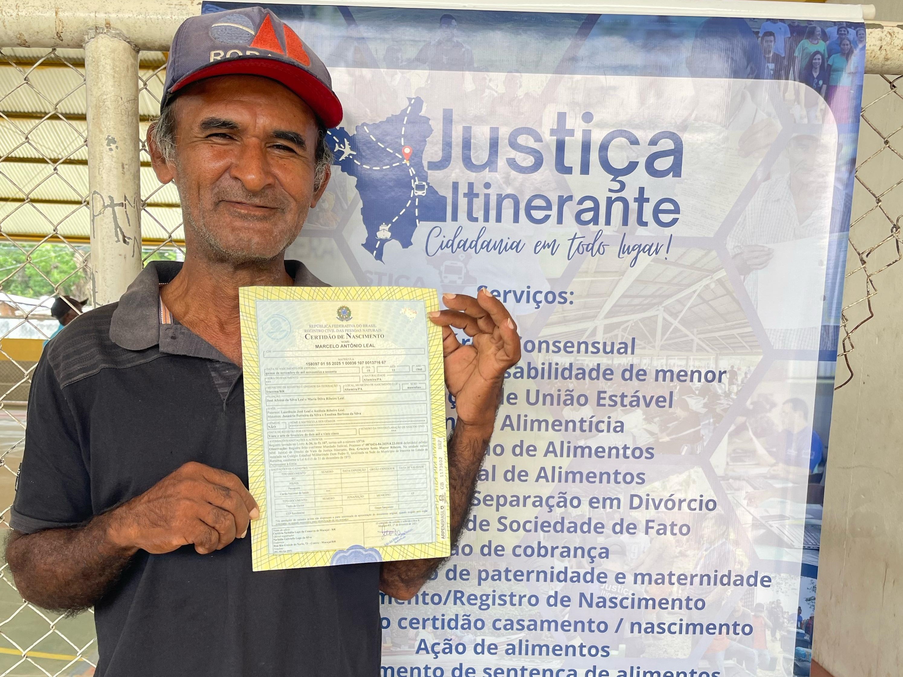  A imagem colorida mostra um homem de pele morena, com barba e bigode, veste uma camiseta cinza escura e um boné vermelho e branco. Ele está em pé, segurando uma certidão de nascimento com as duas mãos, e sorri para a câmera. Ao fundo, há um banner com a inscrição "Justiça Itinerante Cidadania em todo lugar!".