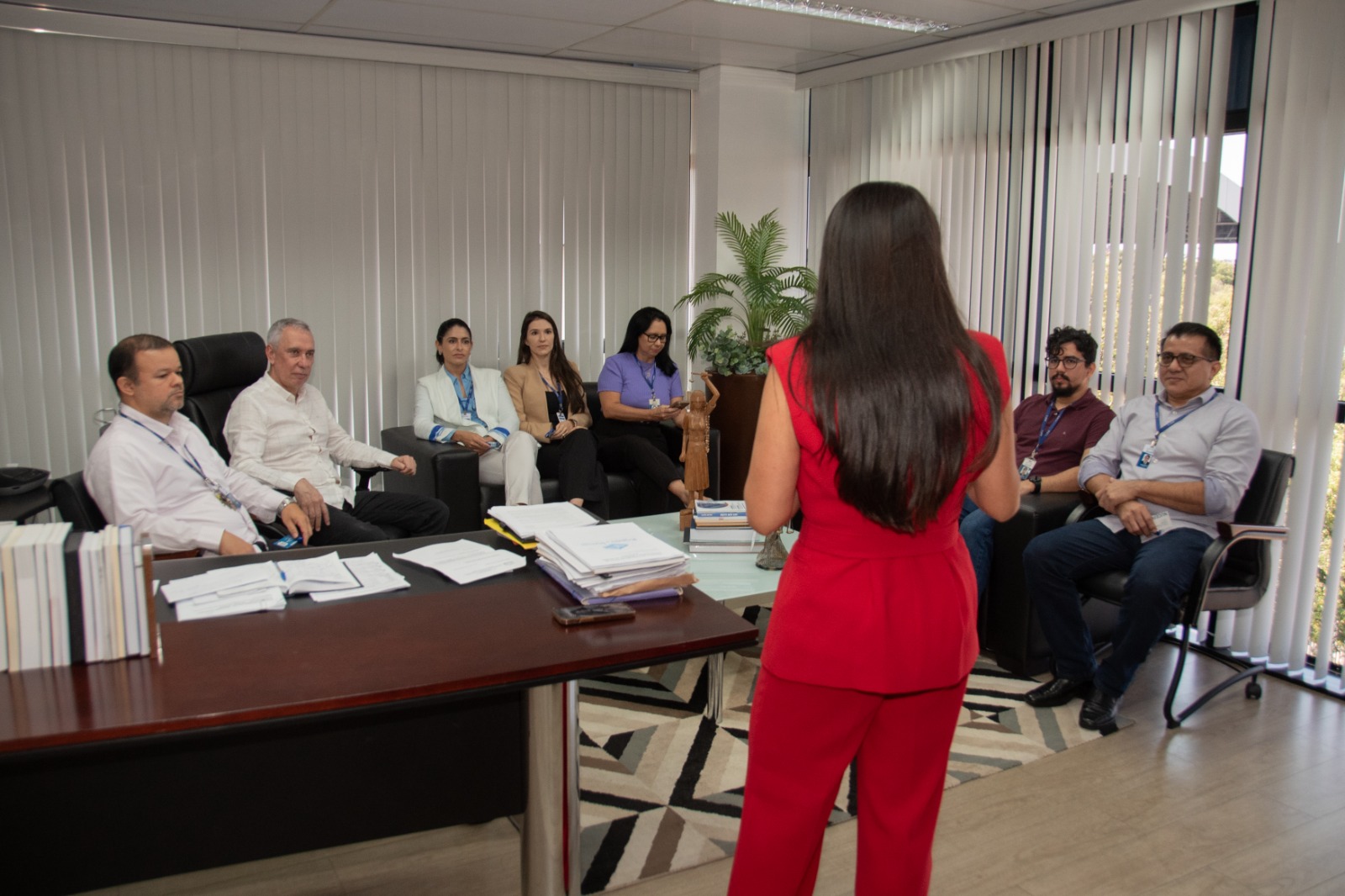 Foto colorida mostra uma sala de gabinete com oito pessoas reunidas. Em primeiro plano, uma mulher de cabelos longos, lisos e escuros, veste conjunto social vermelho e está de costas, falando para o grupo. À esquerda, há uma mesa de madeira com pilhas de documentos e livros. Em frente à mulher, estão sete pessoas sentadas em cadeiras e poltronas: quatro homens e três mulheres, todas vestidas com roupas formais. Foto colorida mostra uma sala de gabinete com oito pessoas reunidas. Em primeiro plano, uma mulher de cabelos longos, lisos e escuros, veste conjunto social vermelho e está de costas, falando para o grupo. À esquerda, há uma mesa de madeira com pilhas de documentos e livros. Em frente à mulher, estão sete pessoas sentadas em cadeiras e poltronas: quatro homens e três mulheres, todas vestidas com roupas formais.