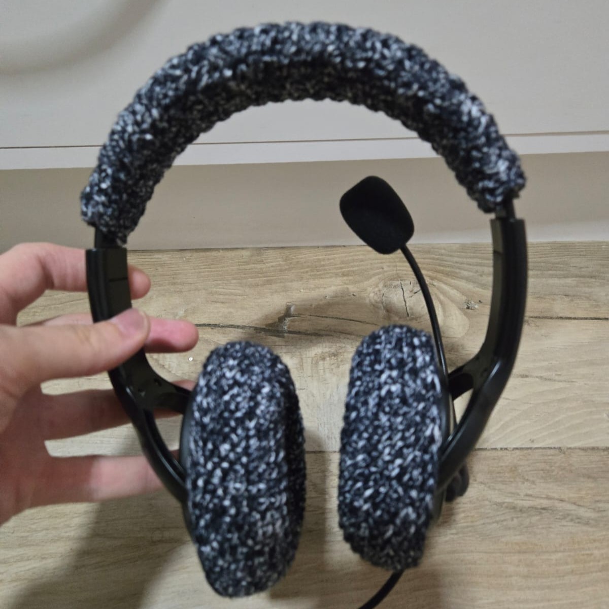Foto colorida mostra uma mão segurando um fone de ouvido grande, com revestimento de crochê preto com branco nas almofadas auriculares.