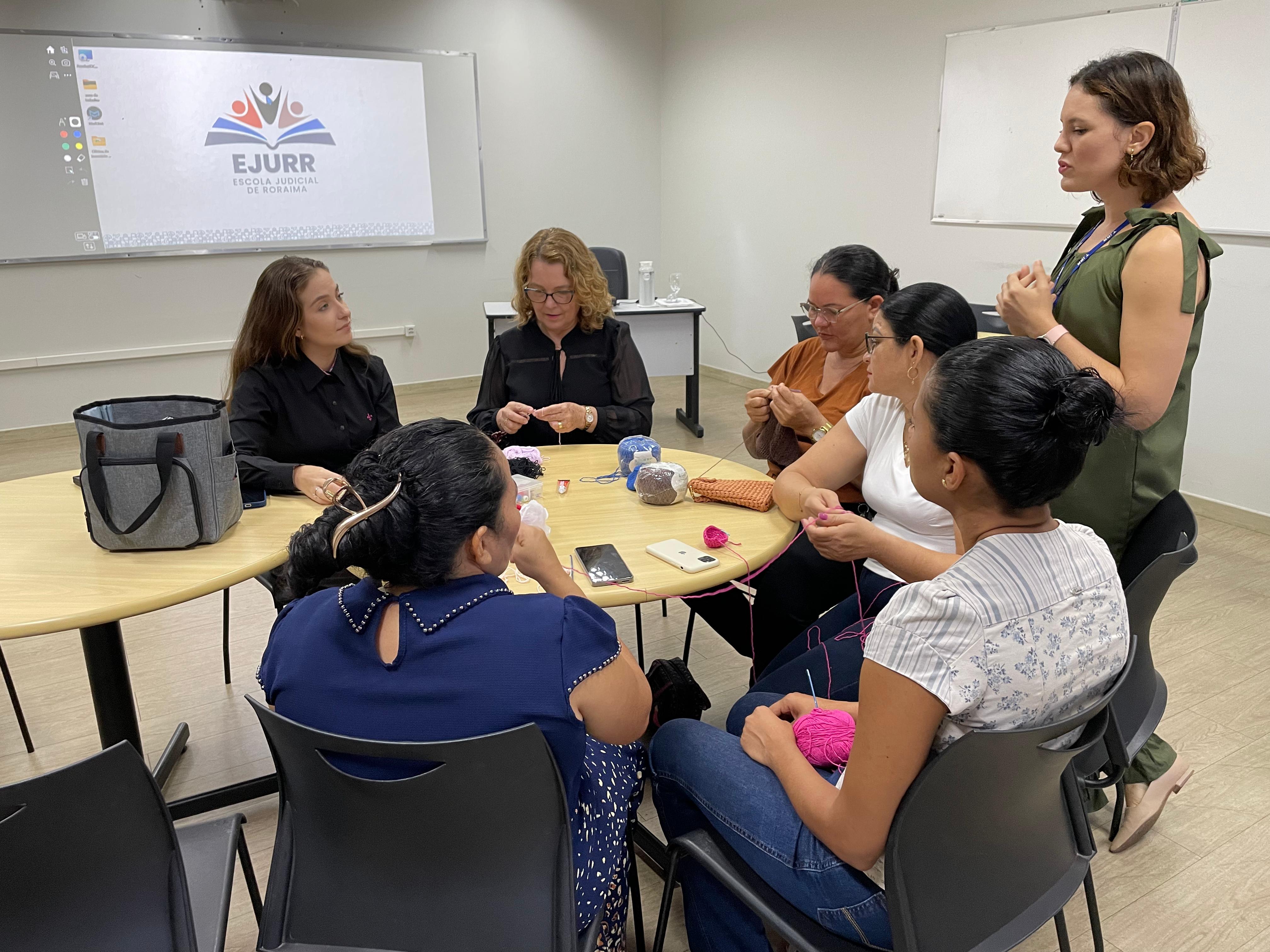 Foto colorida mostra sete mulheres reunidas ao redor de uma mesa oval clara, participando de uma oficina de crochê. 