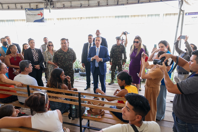 A imagem mostra um grupo de pessoas reunidas embaixo de uma estrutura coberta. Em primeiro plano, algumas pessoas estão sentadas em bancos de madeira, de costas para a câmera, ouvindo atentamente. Em pé, ao centro, um homem de terno azul claro e gravata fala para o público, segurando papéis nas mãos. Ao lado dele, há militares fardados e outras pessoas de terno. À direita, uma fotógrafa registra o momento. No fundo, é possível ver mais pessoas acompanhando a cena.