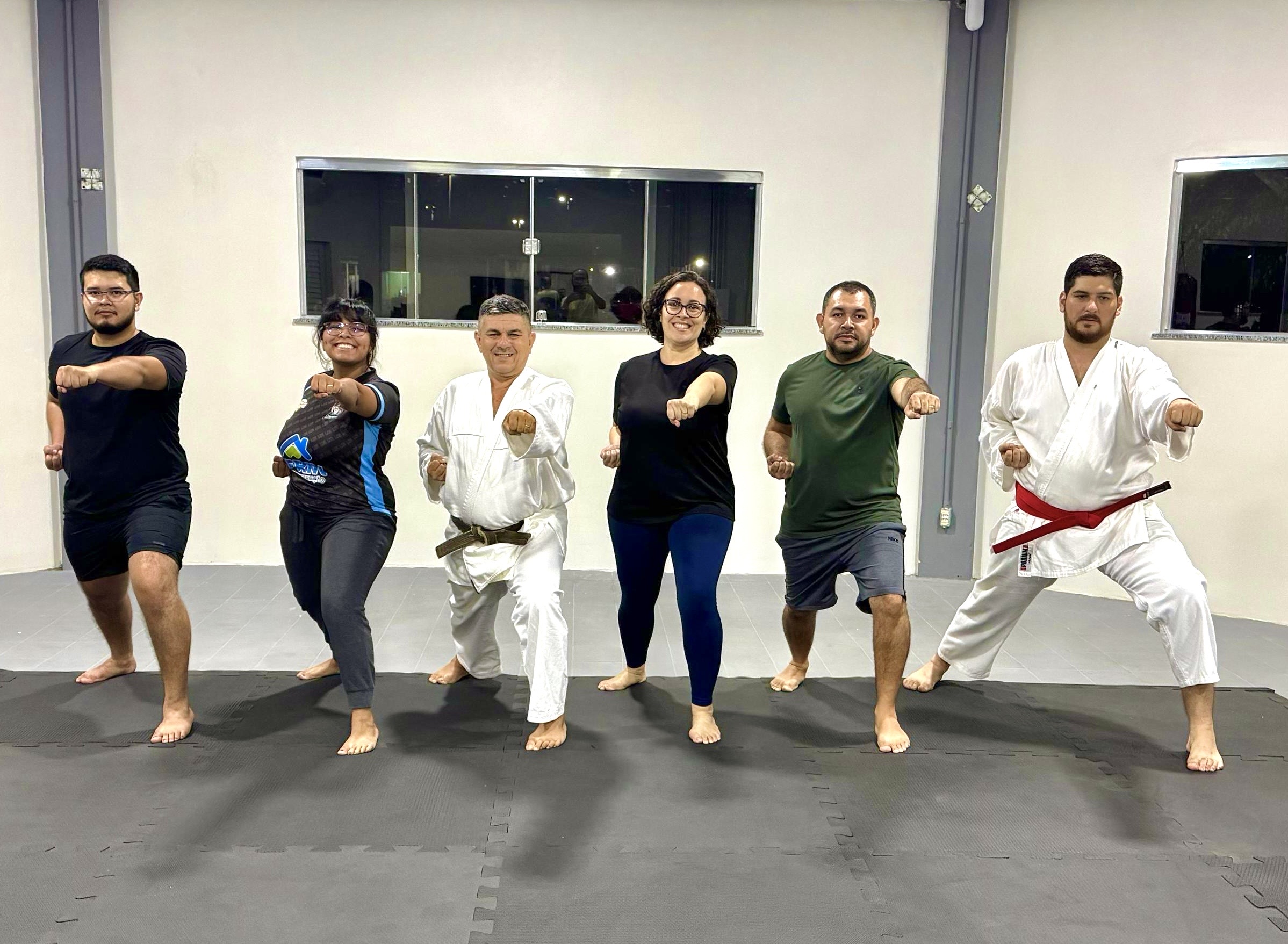 Imagem colorida mostra seis pessoas em uma sala de ginástica. A maioria está descalça, em uma pose de caratê com o punho direito estendido para frente. Um homem está em uma posição de luta com a perna direita dobrada. Todos parecem concentrados. Imagem colorida mostra seis pessoas em uma sala de ginástica. A maioria está descalça, em uma pose de caratê com o punho direito estendido para frente. Um homem está em uma posição de luta com a perna direita dobrada. Todos parecem concentrados.