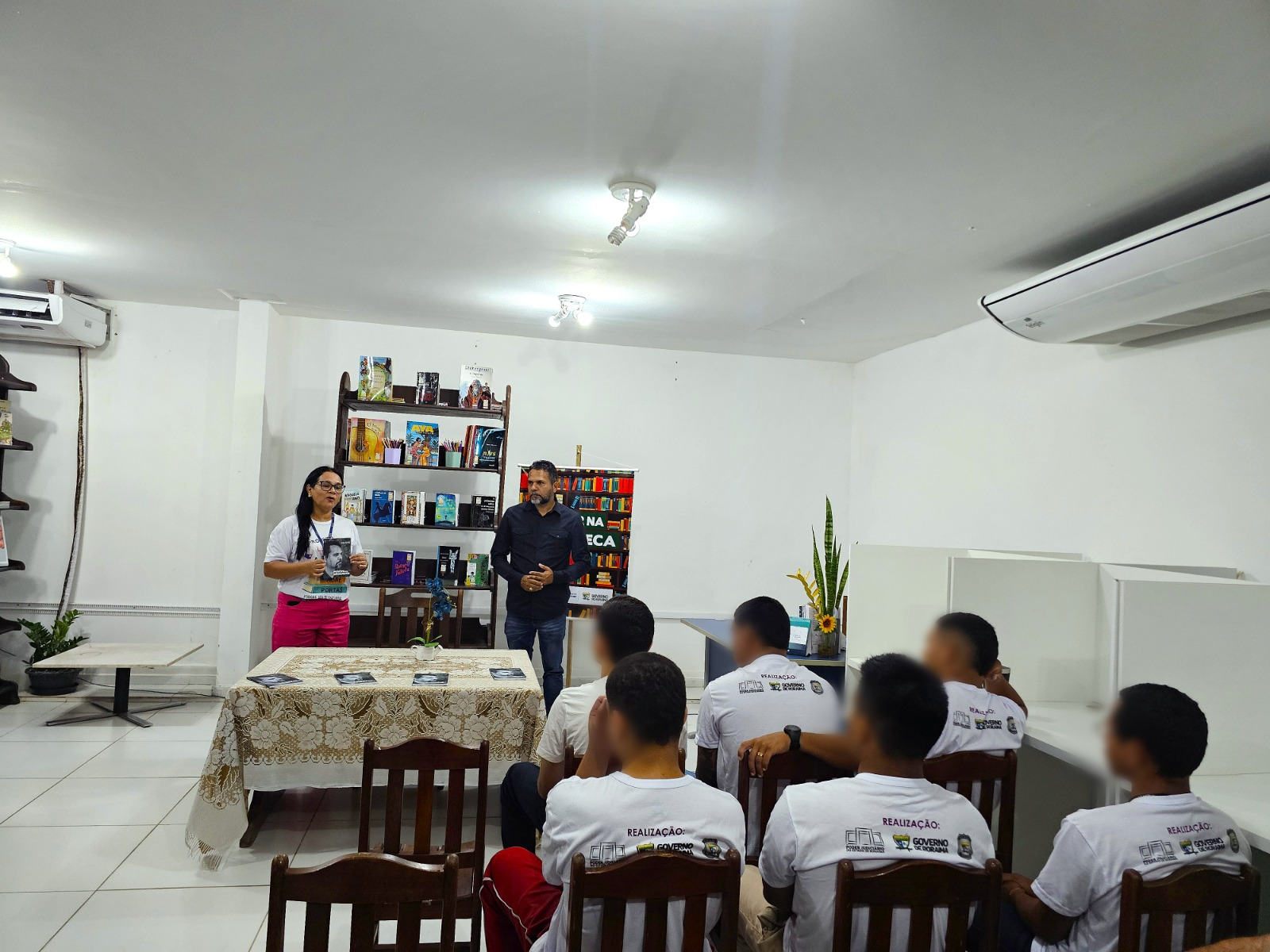 Foto colorida mostra à esquerda, uma mulher de cabelo longo, liso e preto, usando óculos, camiseta branca e calça rosa, segura um livro enquanto fala. Ao lado dela está um homem de camisa preta, barba e cabelos escuros, observando a fala da colega. Ambos estão de pé, à frente de uma mesa coberta por uma toalha rendada bege, sobre a qual há exemplares de livros organizados. Sentados de costas, alguns jovens com camisetas brancas estampadas com os dizeres “Socioeducativo” e logomarcas de instituições parceiras assistem à apresentação. Atrás da dupla, há uma estante de madeira escura com livros coloridos e um banner com a frase “Autor na Biblioteca”. À direita, uma planta com flores amarelas completa a decoração do ambiente.