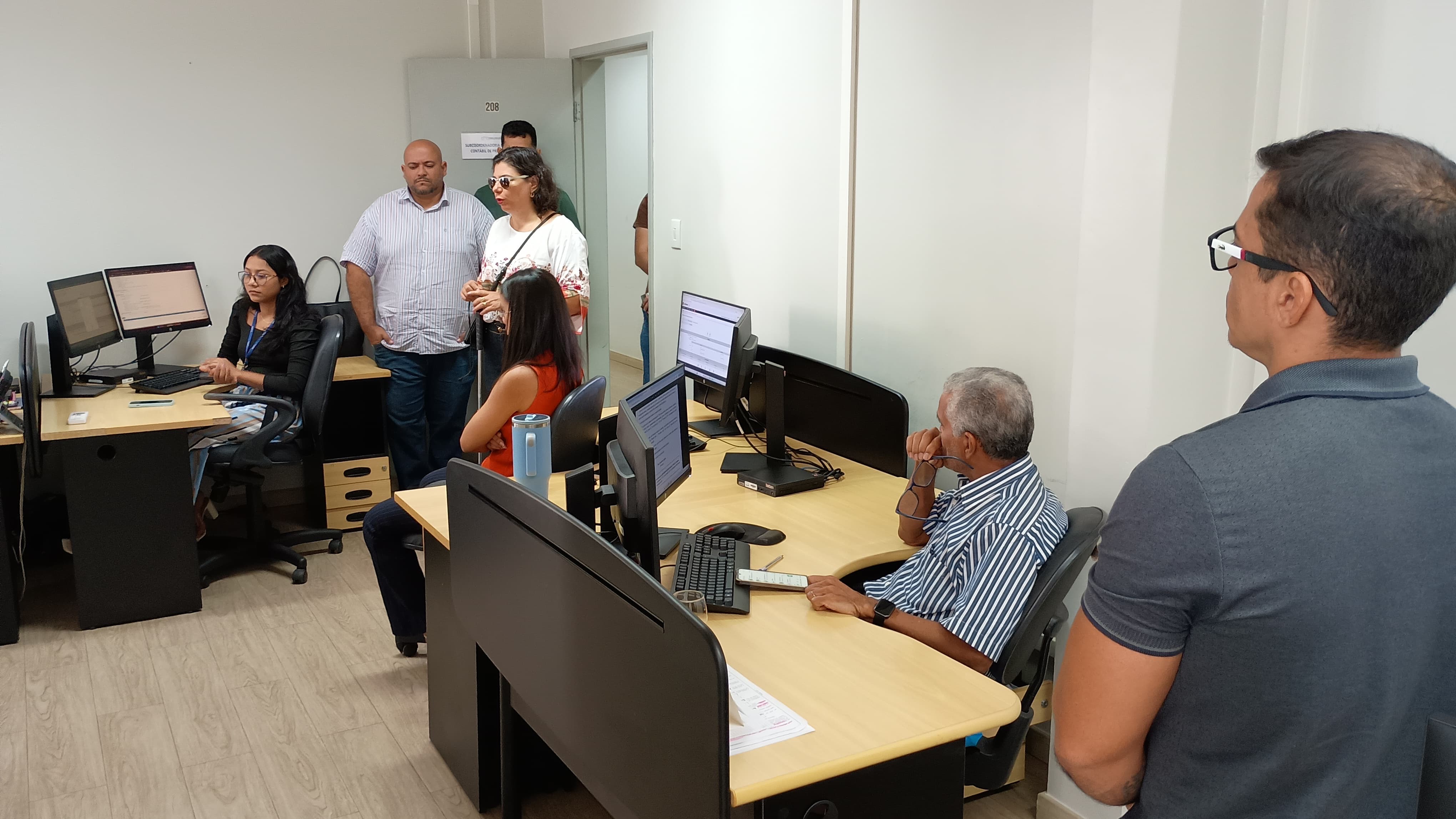 Imagem colorida mostra um escritório onde várias pessoas estão reunidas em pé, olhando para a frente. O ambiente parece ser um escritório com mesas e computadores ao fundo. A maioria das pessoas está vestindo roupas casuais.