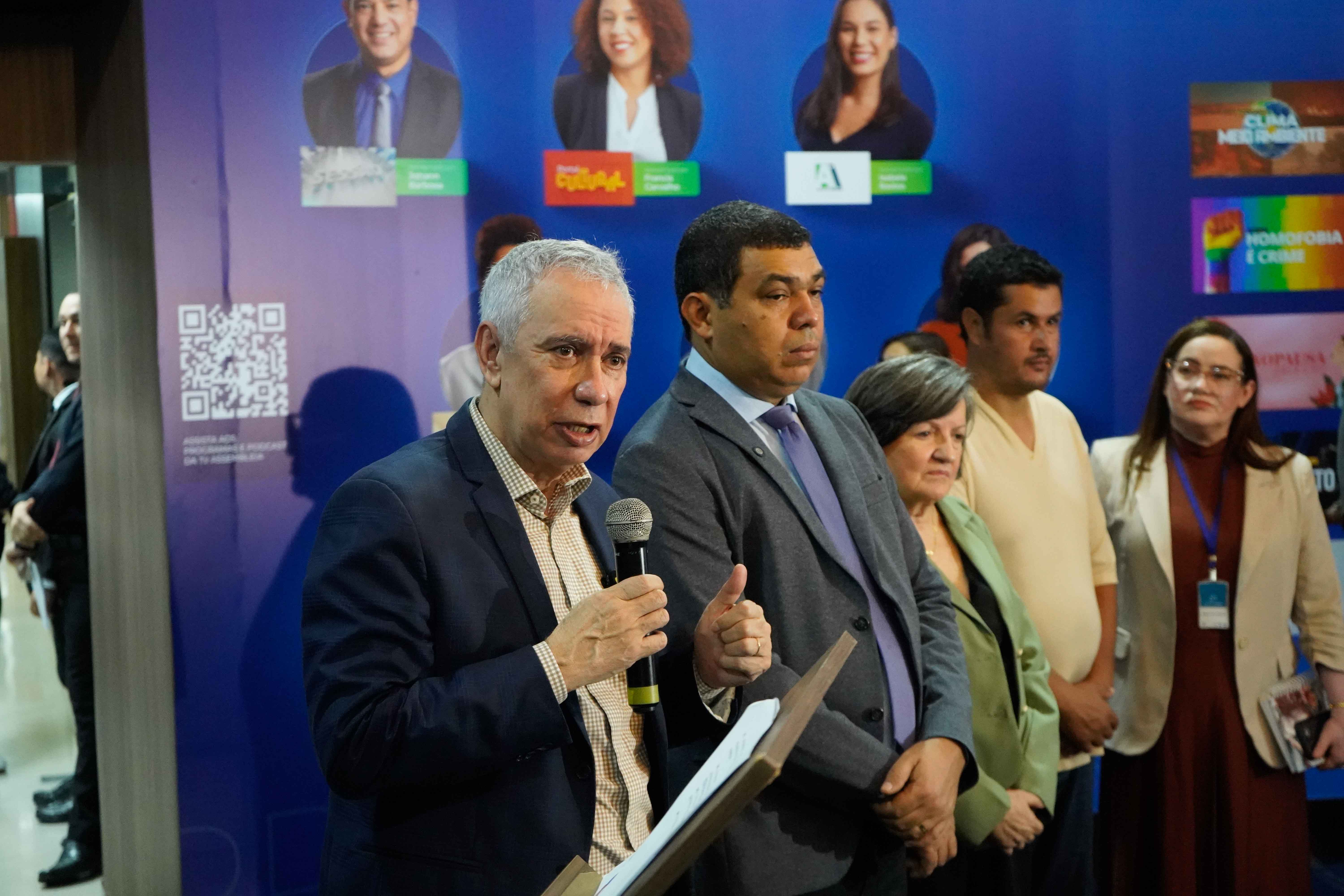 Imagem colorida mostra o Vice-Presidente do Tribunal de Justiça de Roraima, desembargador Almiro Padilha falando no microfone enquanto segura um papel em um púlpito. Ao seu lado, um homem mais jovem de terno cinza faz um gesto com o polegar para cima. Há outras pessoas atrás e ao lado deles.