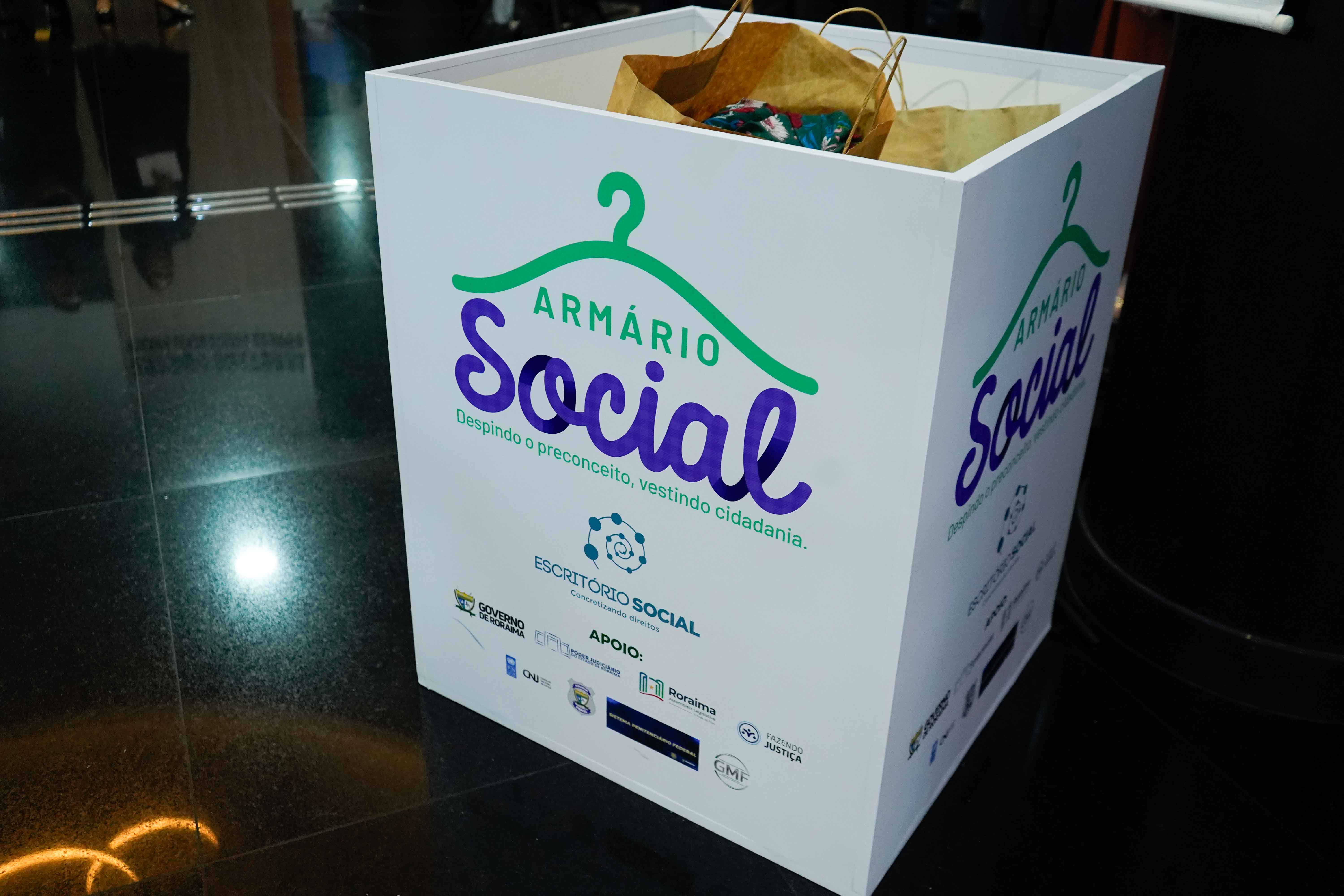Imagem colorida mostra um primeiro plano de uma grande caixa branca no chão, identificada como "Armário Social" com a frase em português "Despindo o preconceito, vestindo cidadania". A caixa contém alguns sacos de papel e pacotes no topo, sugerindo ser um recipiente de doações. Abaixo do nome do projeto, há logotipos de diversas instituições de apoio em pequenas dimensões.