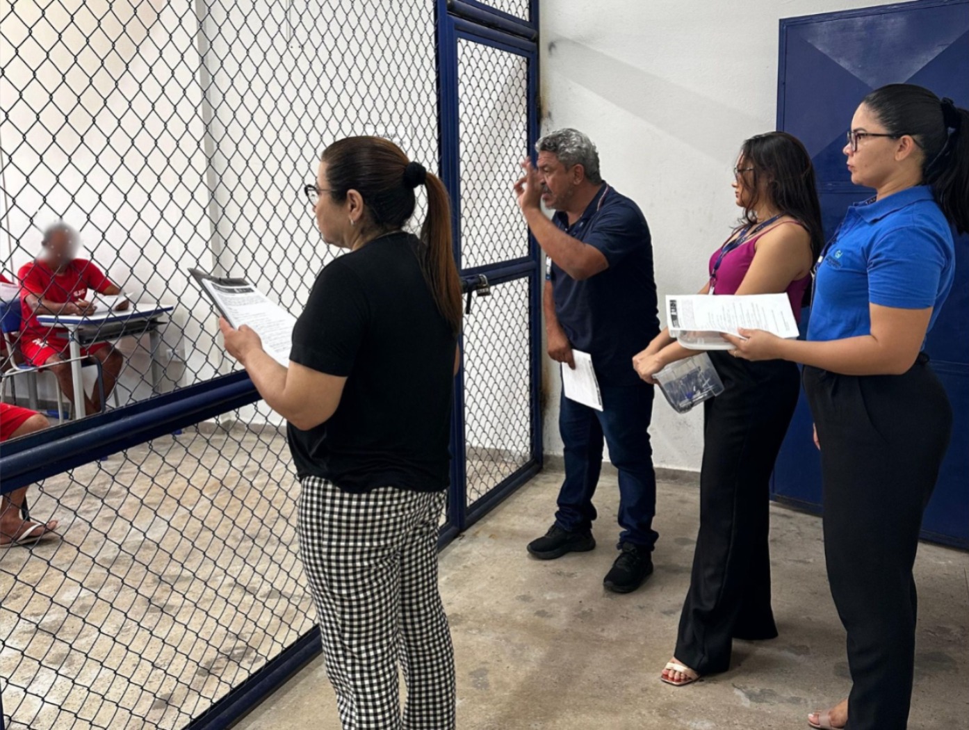 A imagem mostra quatro pessoas posicionadas em frente a uma grade de metal pintada de azul, sendo um homem e três mulheres. Uma das mulheres, de cabelos presos e óculos, segura um questionário; ao seu lado, um homem de barba grisalha faz um gesto com a mão, como se estivesse explicando ou dando orientações. Duas outras mulheres, uma com blusa roxa e outra com camisa azul, também seguram documentos. Do outro lado da grade, presos uniformizados em vermelho estão sentados em carteiras escolares individuais. A imagem mostra quatro pessoas posicionadas em frente a uma grade de metal pintada de azul, sendo um homem e três mulheres. Uma das mulheres, de cabelos presos e óculos, segura um questionário; ao seu lado, um homem de barba grisalha faz um gesto com a mão, como se estivesse explicando ou dando orientações. Duas outras mulheres, uma com blusa roxa e outra com camisa azul, também seguram documentos. Do outro lado da grade, presos uniformizados em vermelho estão sentados em carteiras escolares individuais.
