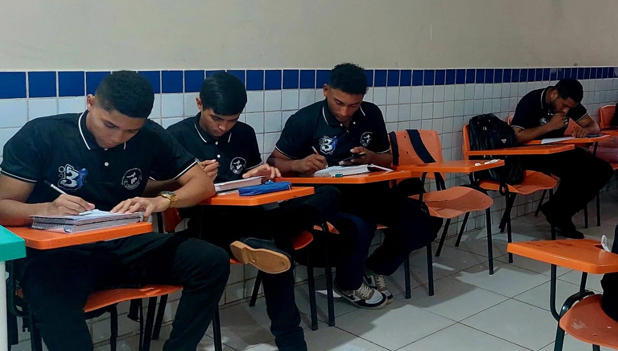 imagem mostra quatro estudantes em uma sala de aula. Eles vestem uniformes pretos com golas e detalhes brancos, que trazem estampados o logotipo da instituição. Sentados em carteiras escolares individuais de cor laranja, respondem questionários aplicados pela pesquisa.  imagem mostra quatro estudantes em uma sala de aula. Eles vestem uniformes pretos com golas e detalhes brancos, que trazem estampados o logotipo da instituição. Sentados em carteiras escolares individuais de cor laranja, respondem questionários aplicados pela pesquisa.