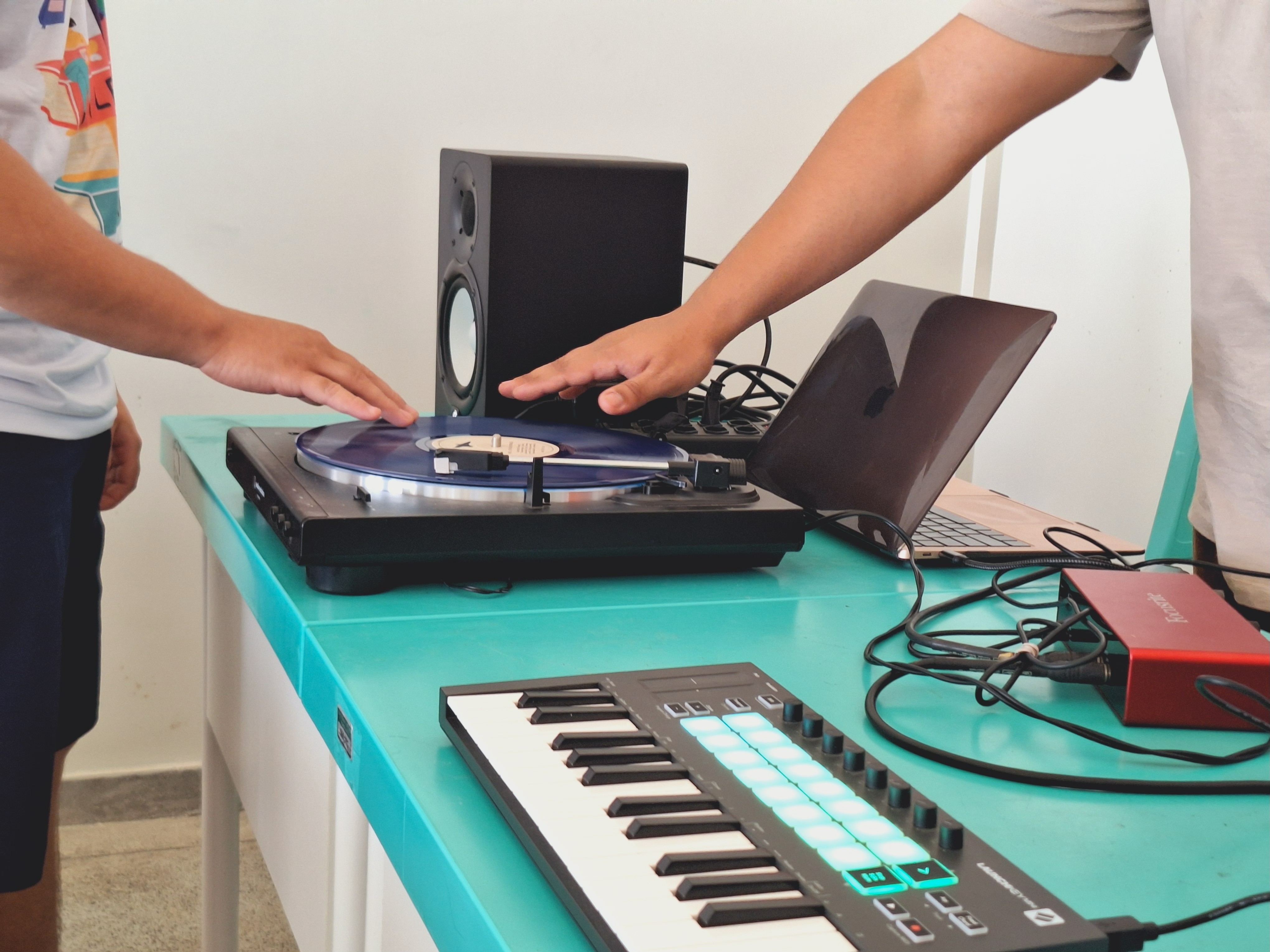 Foto colorida mostra mostra dois jovens interagindo com equipamentos de produção musical sobre uma mesa de cor turquesa. As mãos deles estão posicionadas sobre um toca-discos com um vinil azul. Ao lado, há um teclado com botões iluminados, um laptop aberto, uma caixa de som e uma interface de áudio vermelha com vários cabos conectados.