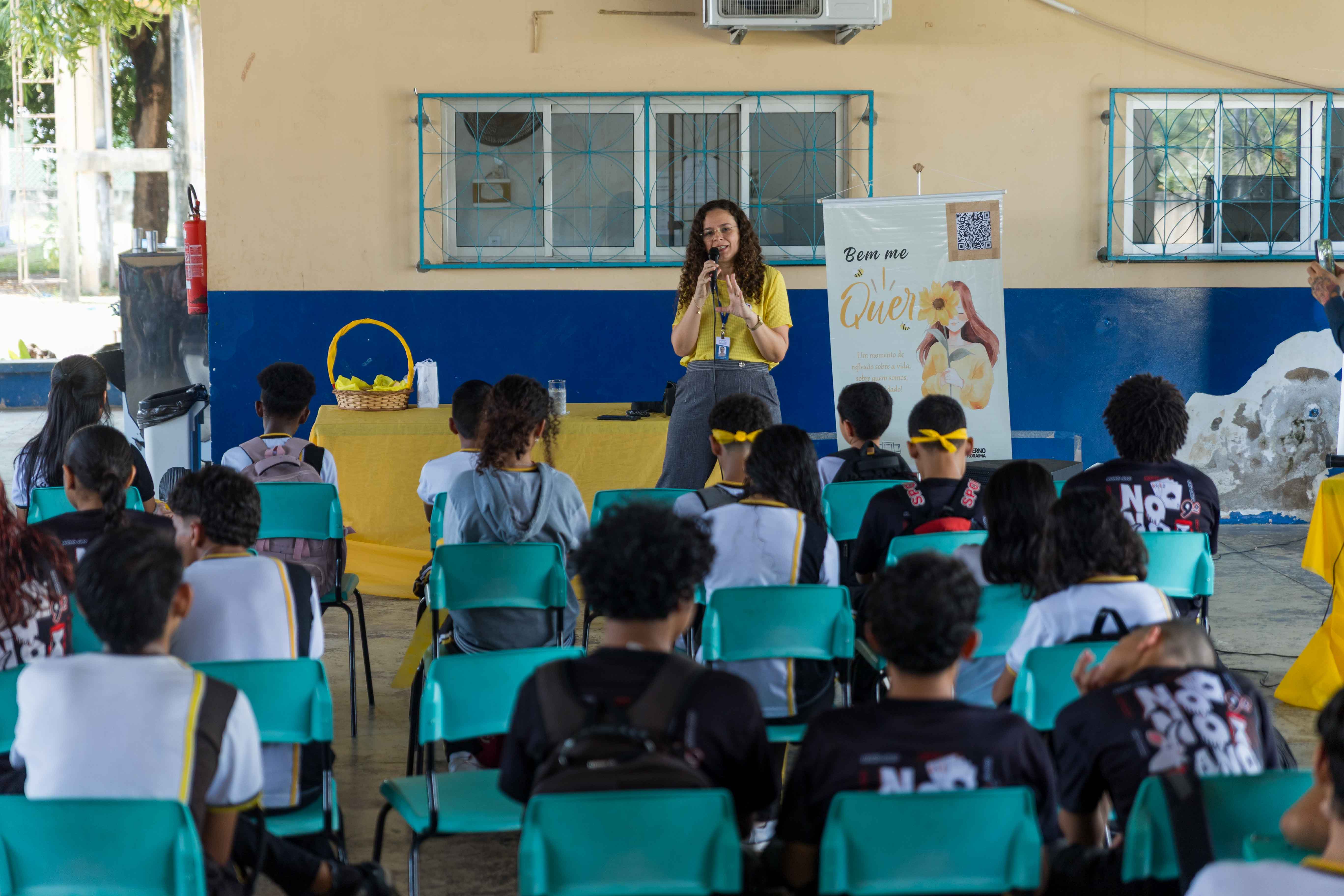 Imagem colorida mostra uma mulher de camisa amarela falando ao microfone para um grupo de jovens sentados em cadeiras azuis. Ao fundo, há um banner amarelo com uma ilustração de girassol e as palavras "Bem me Quero". A cena parece ser uma palestra ou apresentação em um ambiente escolar ou comunitário. Imagem colorida mostra uma mulher de camisa amarela falando ao microfone para um grupo de jovens sentados em cadeiras azuis. Ao fundo, há um banner amarelo com uma ilustração de girassol e as palavras "Bem me Quero". A cena parece ser uma palestra ou apresentação em um ambiente escolar ou comunitário.