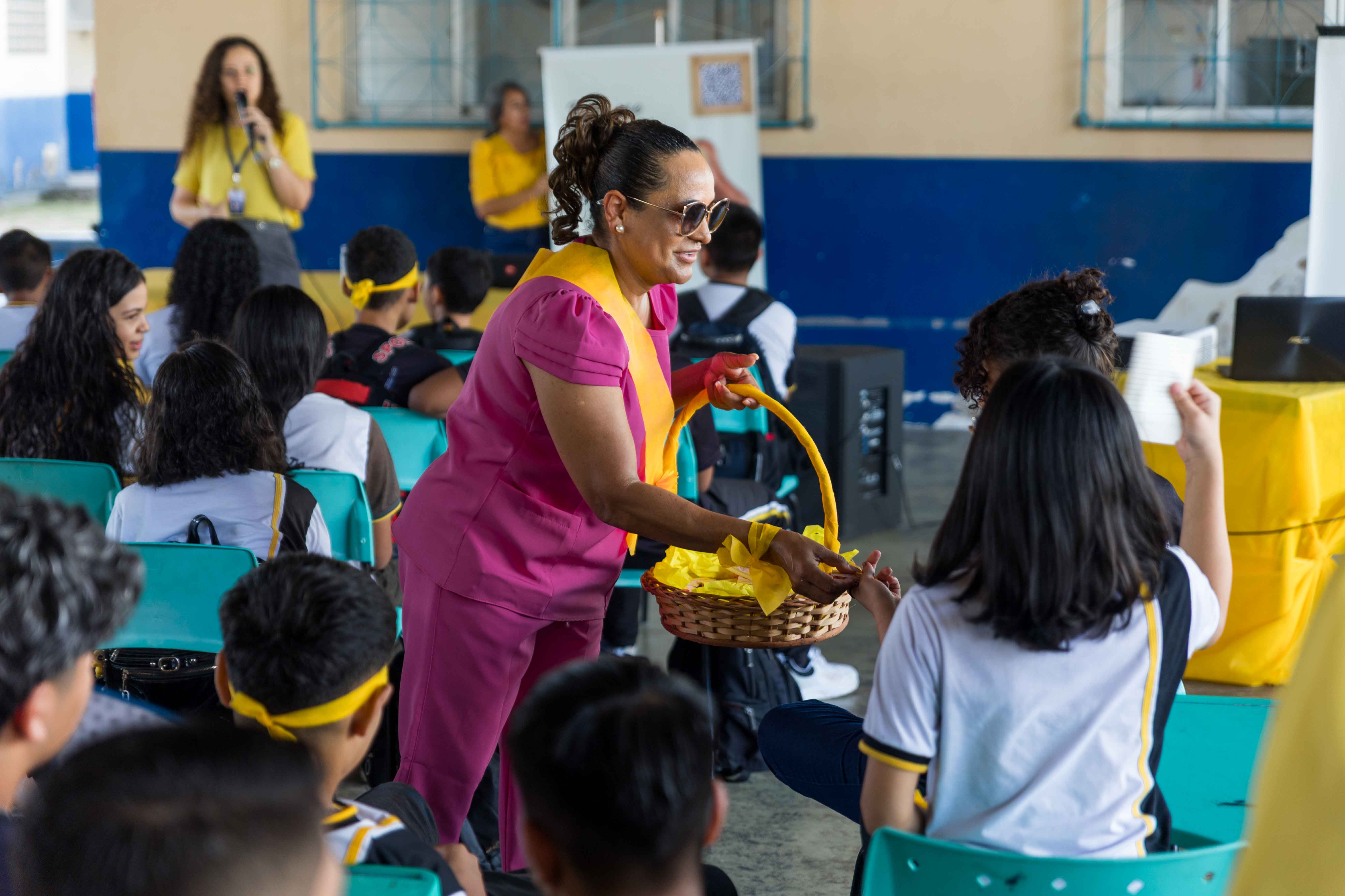 Imagem colorida mostra uma mulher de óculos escuros e uniforme rosa e amarelo, segurando uma cesta de vime com fitas amarelas. Ela se inclina para oferecer algo a uma jovem, enquanto outros estudantes assistem. A cena acontece em um ambiente escolar, com uma mulher ao fundo falando em um microfone. Imagem colorida mostra uma mulher de óculos escuros e uniforme rosa e amarelo, segurando uma cesta de vime com fitas amarelas. Ela se inclina para oferecer algo a uma jovem, enquanto outros estudantes assistem. A cena acontece em um ambiente escolar, com uma mulher ao fundo falando em um microfone.