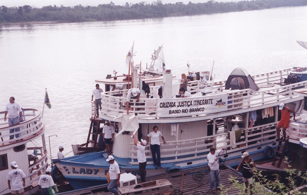 Imagem colorida mostra o barco da Cruzada Justiça Itinerante do Baixo Rio Branco, chegando à comunidade do interior. Na imagem, os tripulantes estão desembarcando.