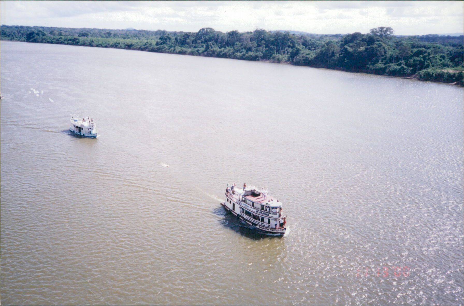 Imagem colorida mostra dois barcos brancos navegando em um rio marrom com margens de floresta. Um barco está à frente do outro.