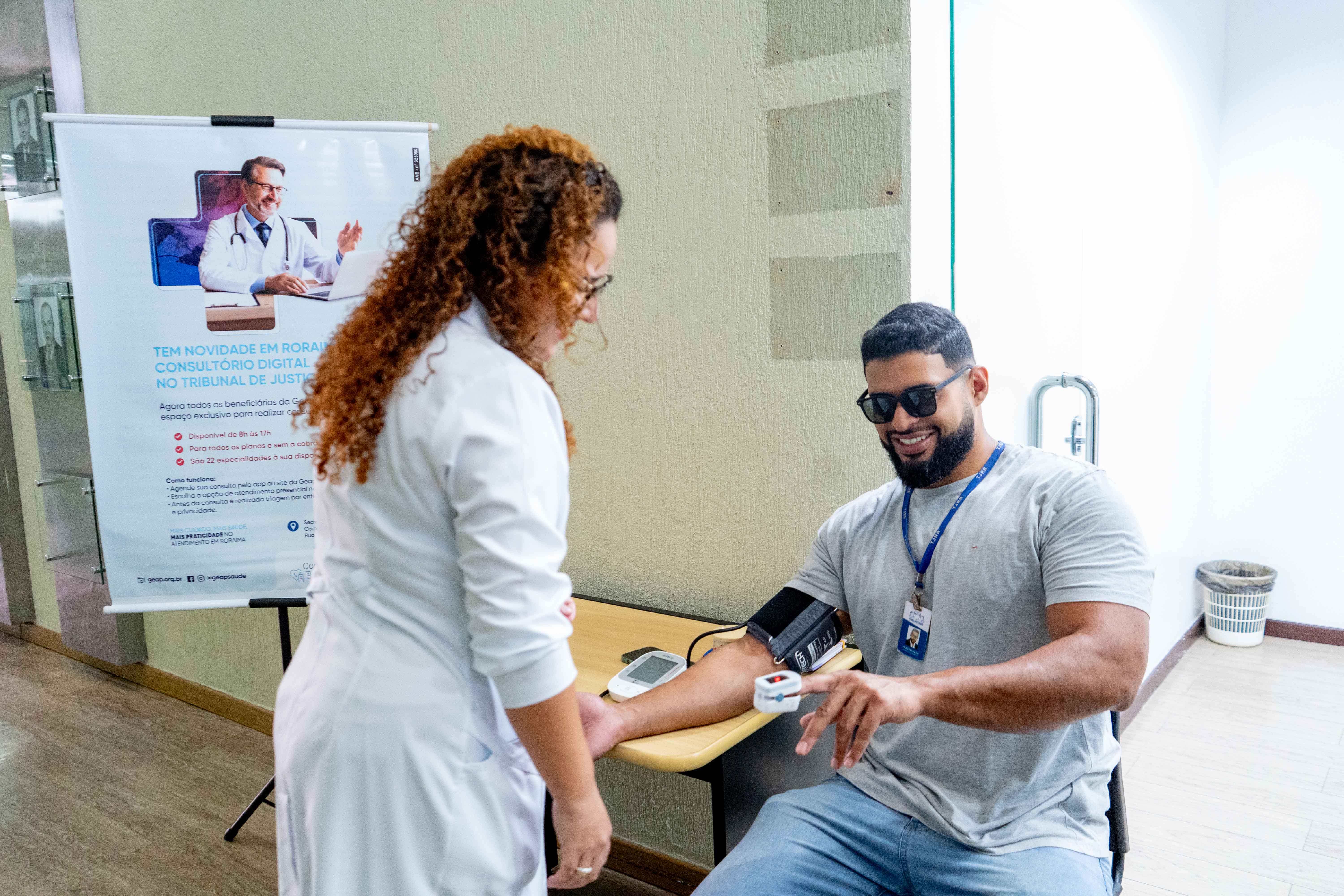 A imagem mostra um ambiente interno onde uma mulher de jaleco branco mede a pressão arterial de um homem com barba, de óculos escuros, que veste uma camisa cinza e está sentado. A mulher está em pé, olhando para o aparelho digital no braço dele, sobre uma mesa de madeira. Ao lado esquerdo, um banner vertical com a imagem de um médico.