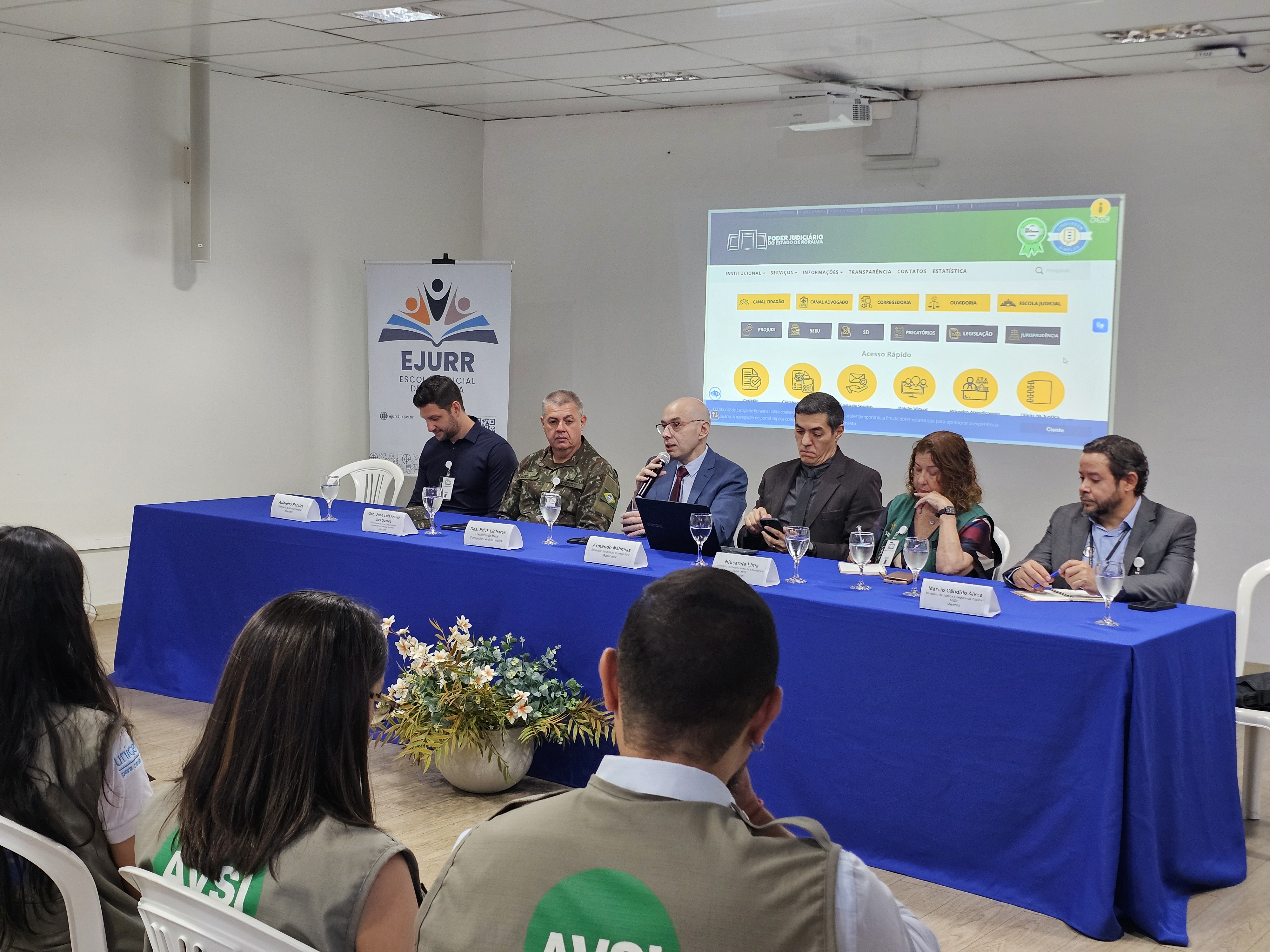 Foto colorida mostra uma mesa solene composta por seis pessoas sentadas, participando de um evento institucional em uma sala da Escola do Poder Judiciário de Roraima.