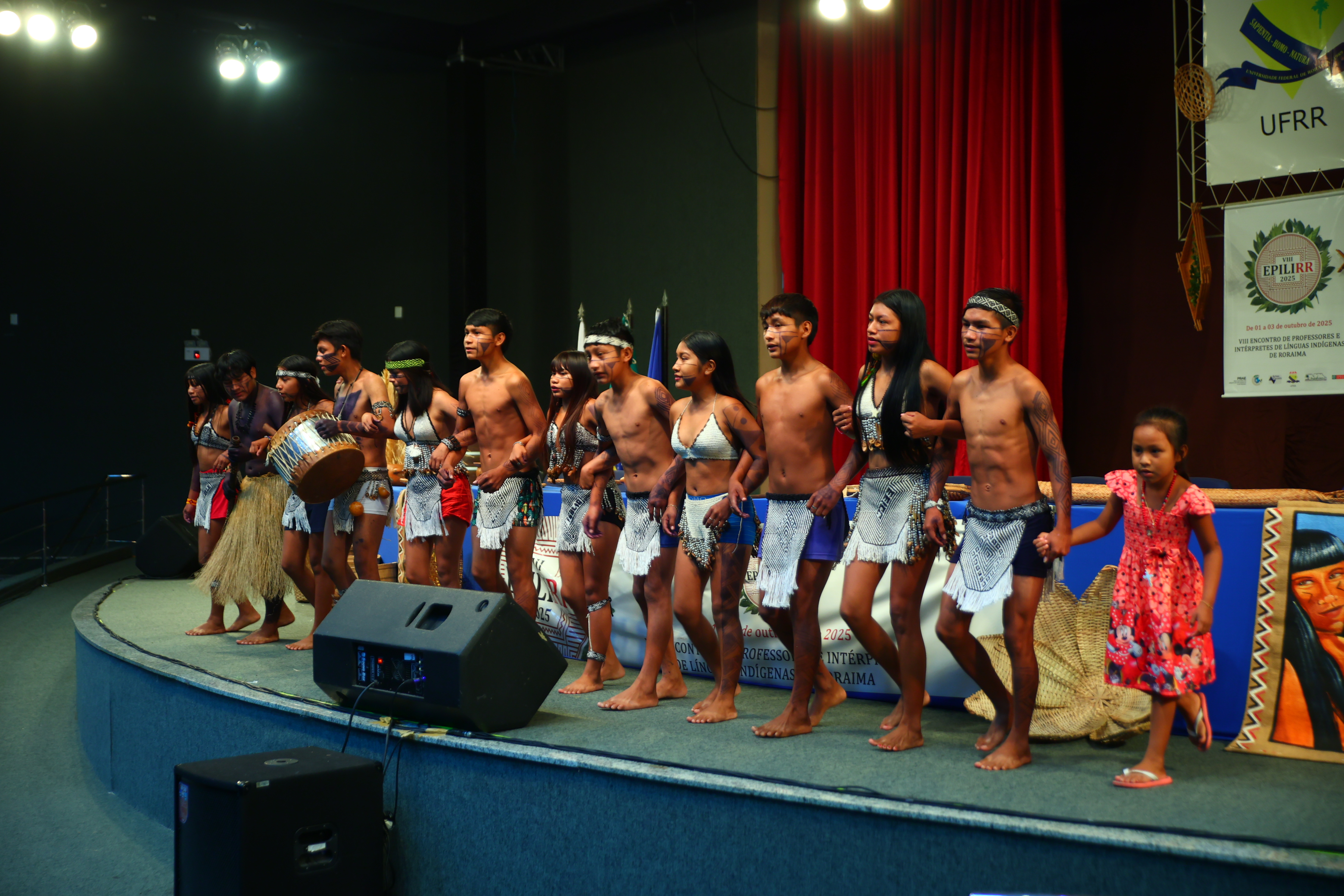 Imagem colorida mostra um grupo de jovens indígenas se apresentando em um palco. Eles usam trajes tradicionais e um carrega um tambor.
