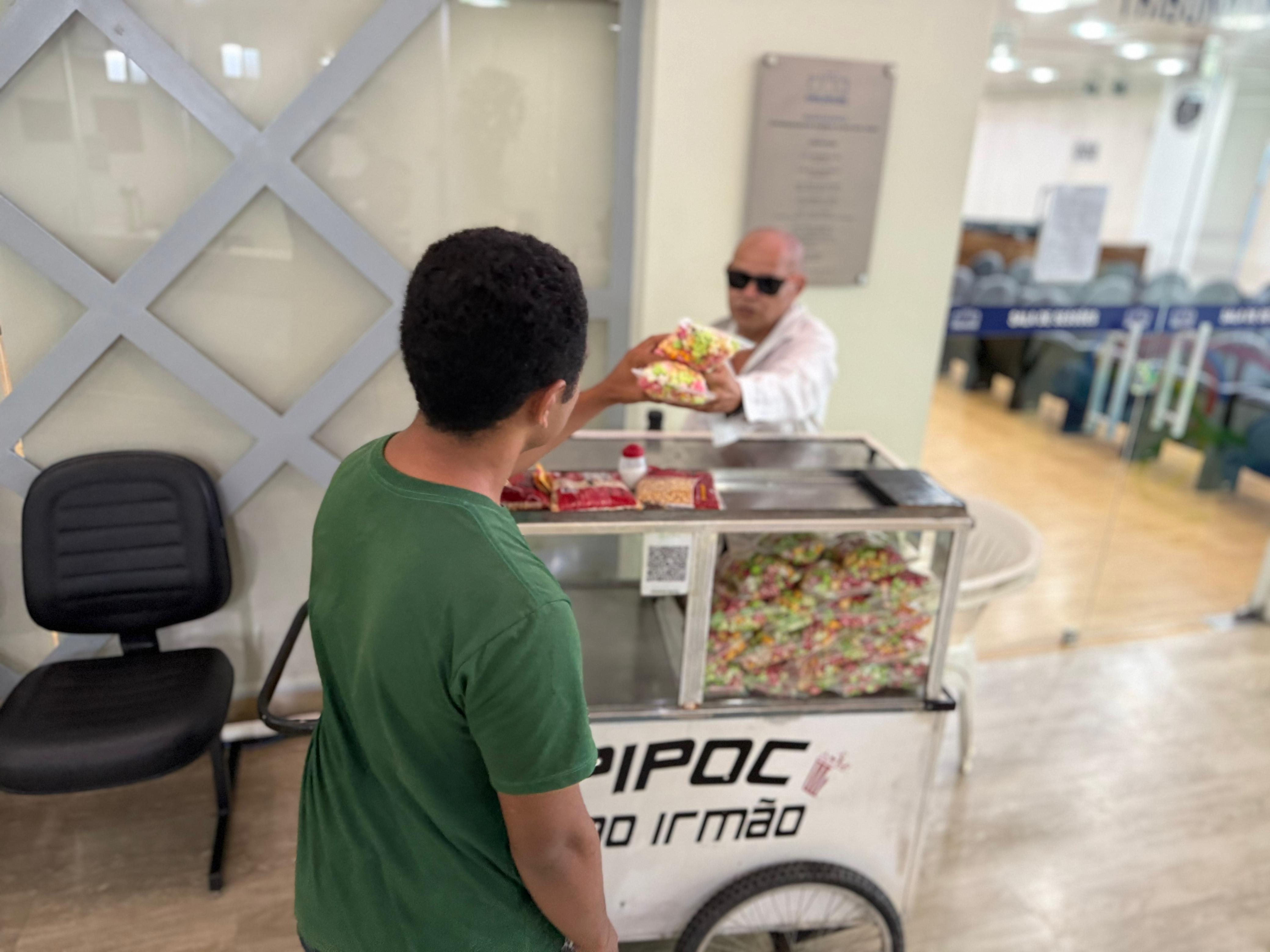 Imagem colorida mostra um carrinho de pipoca sendo operado por um homem de óculos escuros, que está entregando um saco de pipoca colorida para uma pessoa de costas, vestindo uma camiseta verde. O carrinho, de cor branca com rodas e o nome "Pipoca do Irmão" visível na frente, está repleto de sacos de pipoca colorida. A cena se passa em um ambiente interno e iluminado, com uma cadeira escura à esquerda e uma parede com painéis de vidro e metal ao fundo.