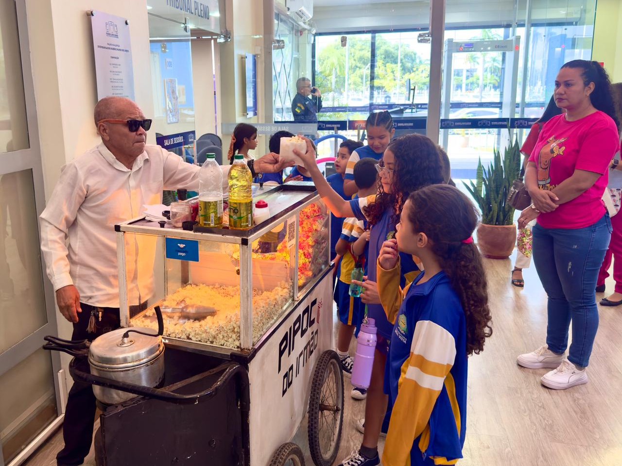 A imagem mostra um hall iluminado, um homem idoso, usando óculos escuros e camisa branca, entrega pipoca em um carrinho. À frente dele, um grupo de crianças com uniforme escolar azul e amarelo forma uma fila, aguardando. Ao lado, uma mulher de camiseta rosa e calça jeans acompanha a cena.