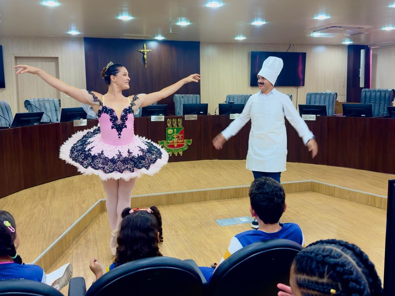 A imagem mostra uma sala ampla com painéis de madeira e cadeiras de reunião, onde uma mulher vestida de bailarina, com figurino rosa e preto, saia rodada e tiara dourada, está com os braços erguidos e um sorriso voltado ao público. Ao lado dela, um homem usa chapéu e avental de chef, também com os braços abertos. À frente, algumas crianças com uniformes azuis e amarelos assistem à apresentação, sentadas em cadeiras estofadas. Ao fundo, o brasão do Tribunal de Justiça de Roraima está visível na parede, sob uma cruz dourada