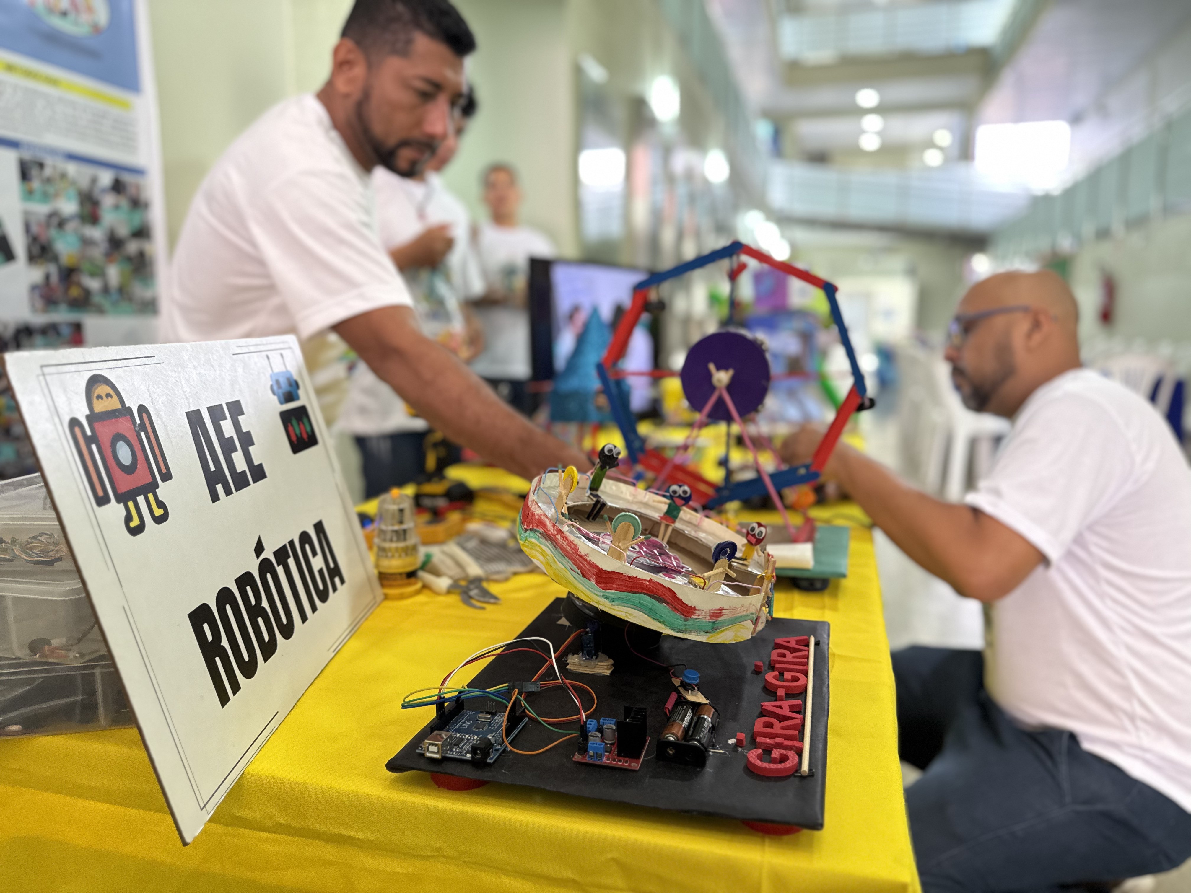 A imagem mostra um corredor iluminado, onde duas pessoas montam e ajustam maquetes coloridas sobre uma mesa coberta com toalha amarela. À frente, um cartaz exibe o título “AEE Robótica”, acompanhado de desenhos de robôs. Na mesa, há projetos feitos com materiais recicláveis, fios, motores e componentes eletrônicos, incluindo uma maquete chamada “Gira-Gira”, com pequenos bonecos e peças móveis. Um homem de barba, vestindo camiseta branca, trabalha em um dos modelos, enquanto outro homem faz ajustes em outro protótipo.