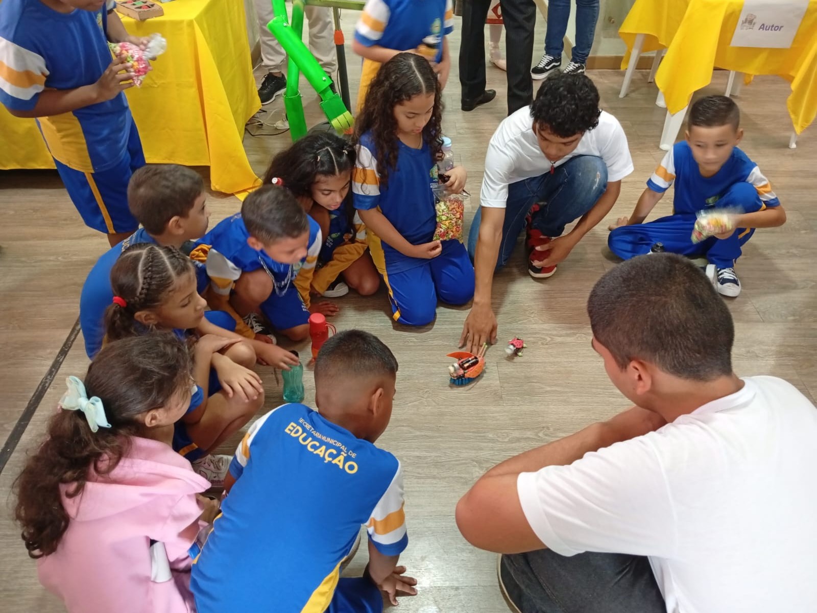 A imagem mostra um grupo de crianças, vestidas com uniformes escolares azuis e amarelos, sentado em círculo no chão, observando atentamente pequenos robôs em movimento. Dois jovens, de camiseta branca, demonstram os brinquedos robóticos, posicionando-os no centro do grupo. Ao fundo, há mesas cobertas com toalhas amarelas e pessoas circulando pelo ambiente.