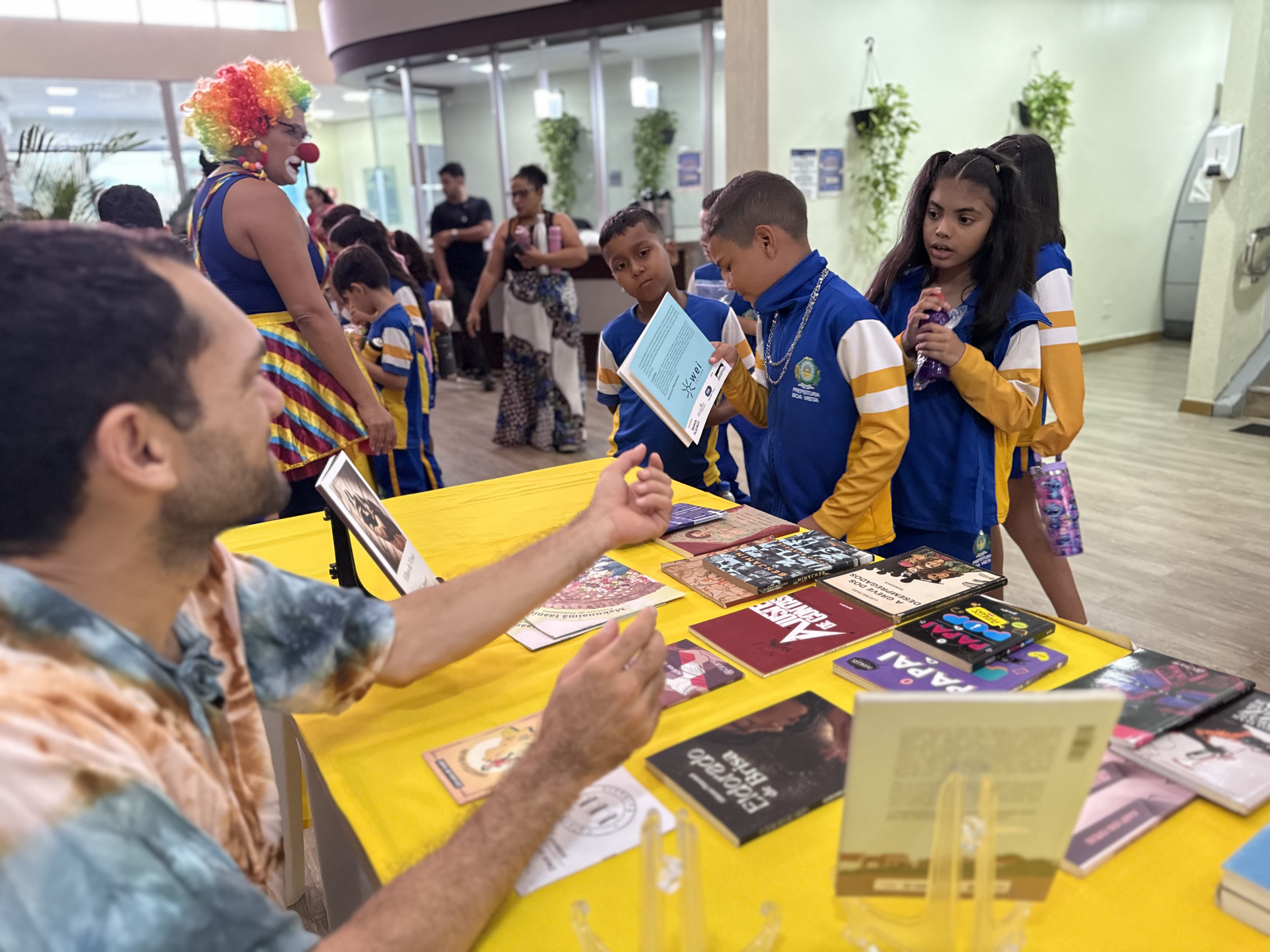A imagem mostra um ambiente interno, um homem sentado atrás de uma mesa coberta com toalha amarela conversa com crianças. Sobre a mesa, há vários livros expostos, com capas coloridas e títulos diversos. Uma menina segura um dos livros e o mostra para um colega, enquanto outras observam com curiosidade. As crianças vestem uniformes escolares azuis e amarelos e carregam garrafinhas de água. Ao fundo, uma pessoa vestida de palhaço, com peruca e roupa colorida, interage com outros alunos.
