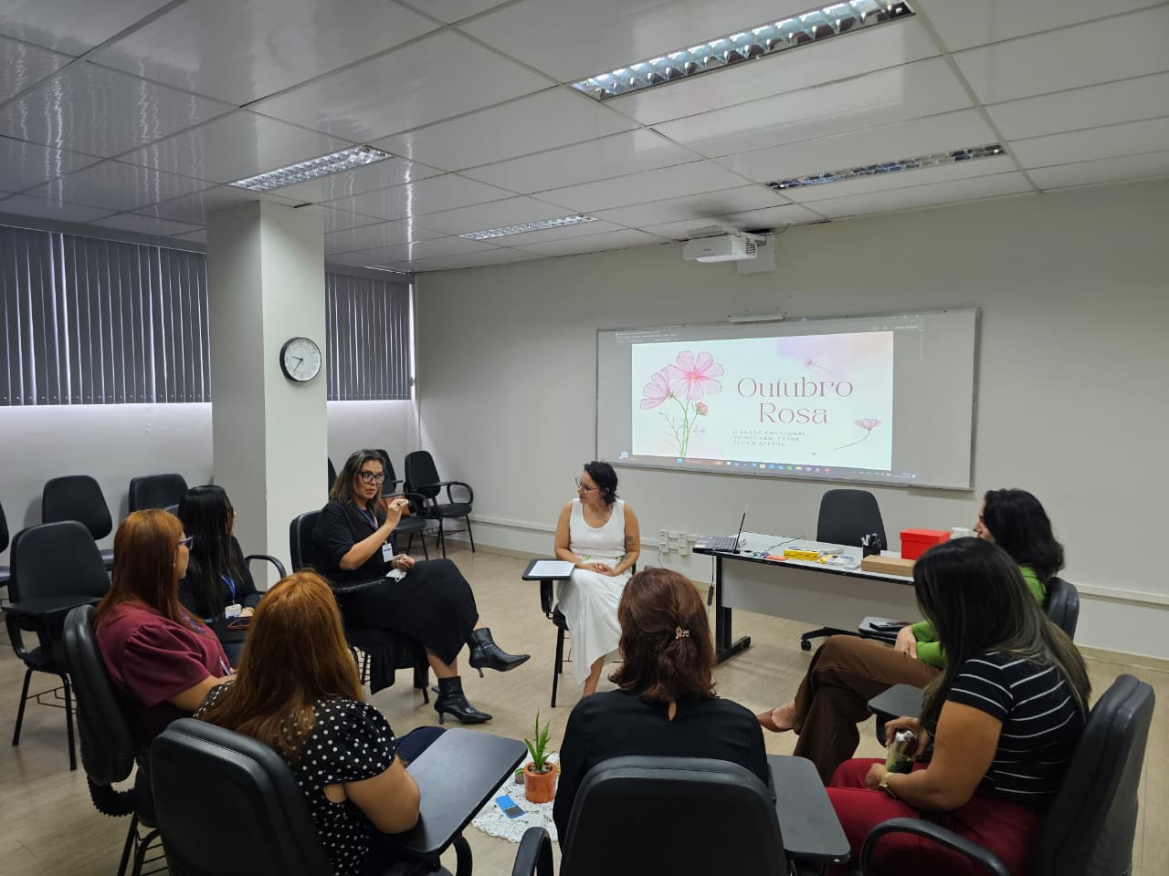 A imagem colorida mostra uma roda de conversa com nove mulheres, reunidas em uma sala de reuniões clara e organizada. As cadeiras estão dispostas em círculo, promovendo um ambiente de diálogo acolhedor. No centro, uma mulher de vestido branco comanda a conversa. A imagem colorida mostra uma roda de conversa com nove mulheres, reunidas em uma sala de reuniões clara e organizada. As cadeiras estão dispostas em círculo, promovendo um ambiente de diálogo acolhedor. No centro, uma mulher de vestido branco comanda a conversa.