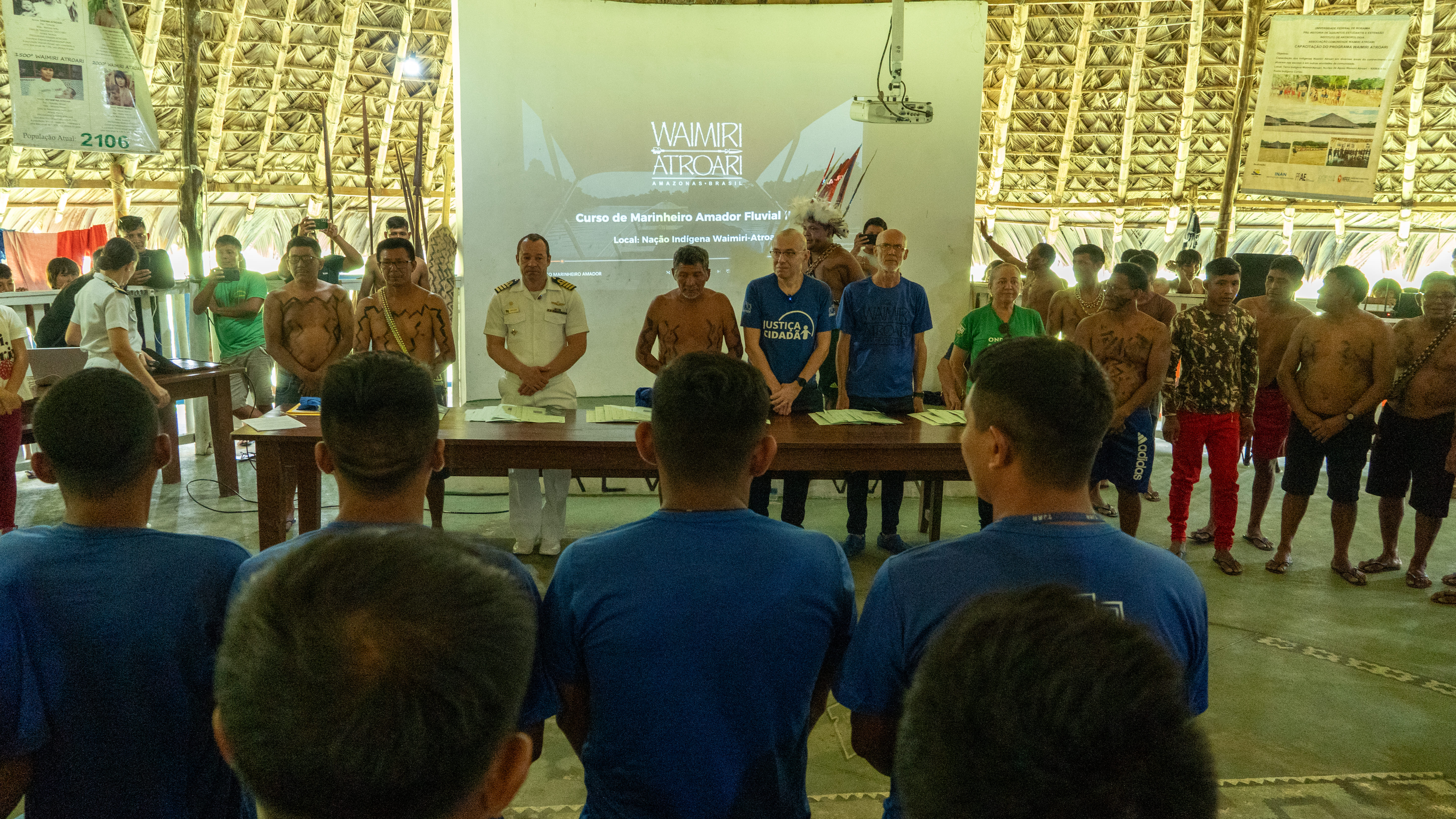 Participantes indígenas e autoridades estão perfilados em frente a uma mesa, durante a abertura de um curso de Marinha Fluvial na Nação Indígena Waimiri-Atroari. Um telão ao fundo exibe o nome do curso e da comunidade.