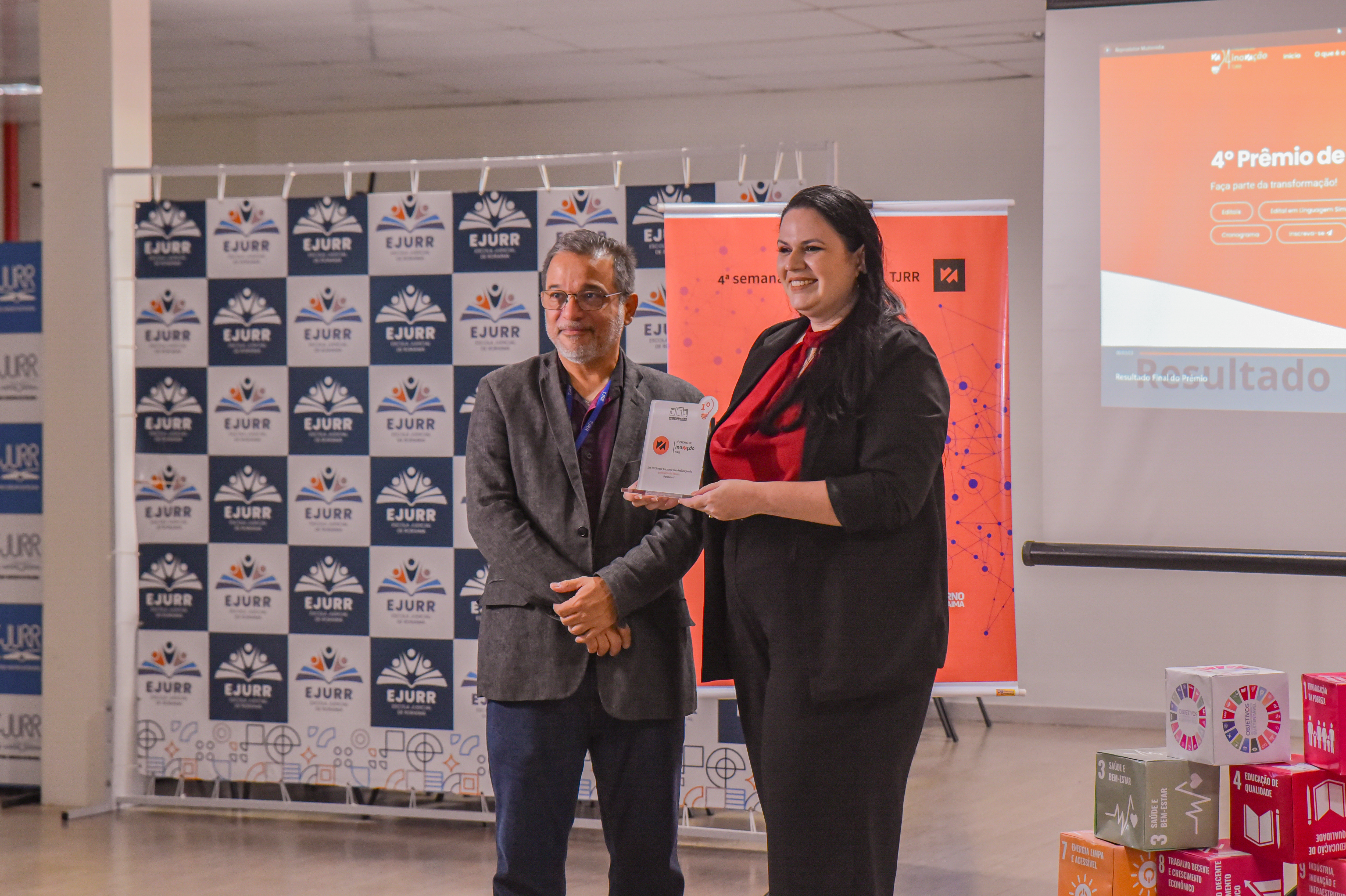Imagem colorida mostra dois participantes em uma cerimônia de premiação na área interna, com fundo de painel institucional da EJURR e banners da Semana de Inovação do TJRR. À direita, segurando o troféu, está Ariane Hayana, integrante da equipe vencedora do 4º Prêmio de Inovação do TJRR, vestida com blusa vermelha e blazer preto. À esquerda, está o secretário-geral do TJRR, Hermenegildo D’Ávila, usando blazer cinza. Ambos posam sorrindo e o ambiente indica um momento de celebração formal. Imagem colorida mostra dois participantes em uma cerimônia de premiação na área interna, com fundo de painel institucional da EJURR e banners da Semana de Inovação do TJRR. À direita, segurando o troféu, está Ariane Hayana, integrante da equipe vencedora do 4º Prêmio de Inovação do TJRR, vestida com blusa vermelha e blazer preto. À esquerda, está o secretário-geral do TJRR, Hermenegildo D’Ávila, usando blazer cinza. Ambos posam sorrindo e o ambiente indica um momento de celebração formal.