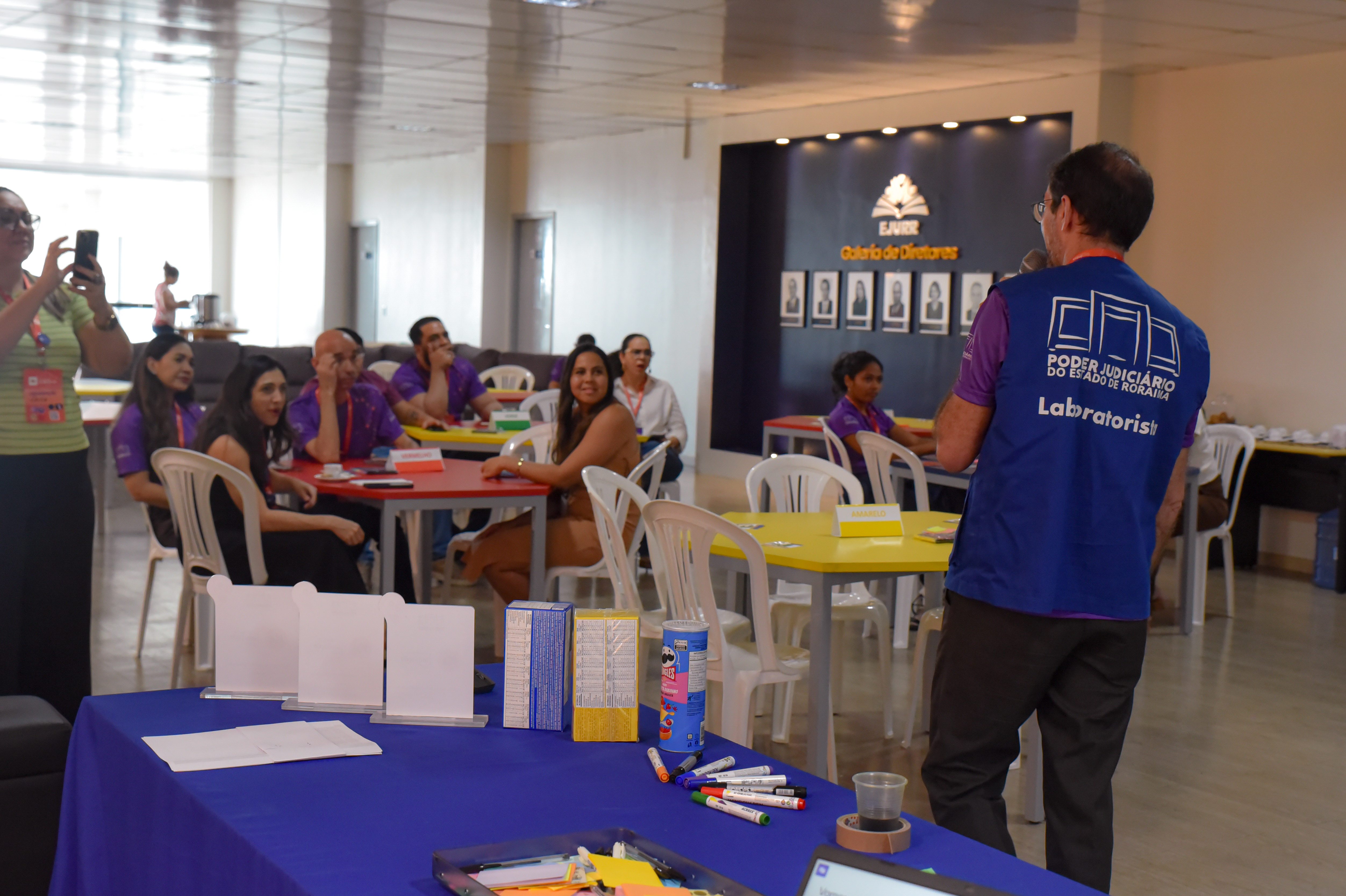 Imagem colorida mostra um ambiente de oficina ou dinâmica coletiva, com mesas coloridas e pessoas sentadas, atentas a um facilitador. Quem está apresentando é France James, chefe do Laboratório de Inovação do TJRR, de costas para a câmera, vestindo um colete azul com a inscrição “Poder Judiciário do Estado de Roraima - Laboratorista”. Diversos materiais de papelaria estão sobre a mesa à frente e, ao fundo, participantes observam e interagem, alguns usando camisetas roxas do evento. O clima é descontraído e colaborativo Imagem colorida mostra um ambiente de oficina ou dinâmica coletiva, com mesas coloridas e pessoas sentadas, atentas a um facilitador. Quem está apresentando é France James, chefe do Laboratório de Inovação do TJRR, de costas para a câmera, vestindo um colete azul com a inscrição “Poder Judiciário do Estado de Roraima - Laboratorista”. Diversos materiais de papelaria estão sobre a mesa à frente e, ao fundo, participantes observam e interagem, alguns usando camisetas roxas do evento. O clima é descontraído e colaborativo