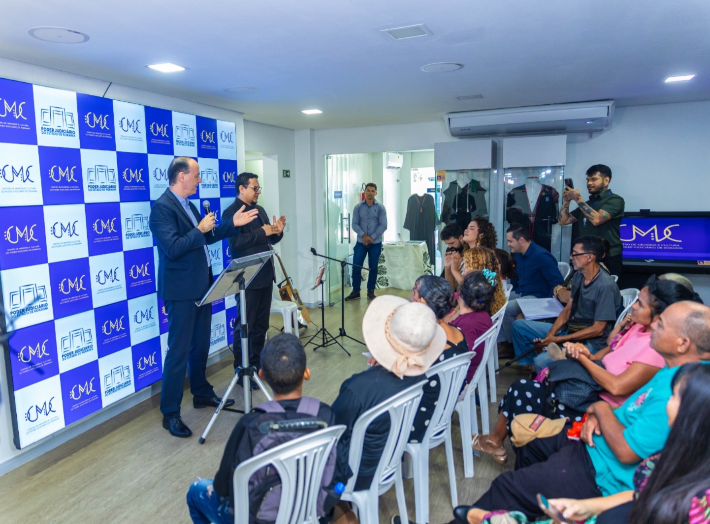 Uma apresentação ou discurso está ocorrendo em um espaço interno. Dois homens estão em frente a um backdrop azul e branco com um logo que inclui as letras "CMC" (e possivelmente "Roraima" ou outro texto menor), falando para uma plateia sentada em cadeiras brancas. Há equipamentos de áudio (microfones) e vídeo (uma TV lateral exibindo o mesmo logo). Uma apresentação ou discurso está ocorrendo em um espaço interno. Dois homens estão em frente a um backdrop azul e branco com um logo que inclui as letras "CMC" (e possivelmente "Roraima" ou outro texto menor), falando para uma plateia sentada em cadeiras brancas. Há equipamentos de áudio (microfones) e vídeo (uma TV lateral exibindo o mesmo logo).
