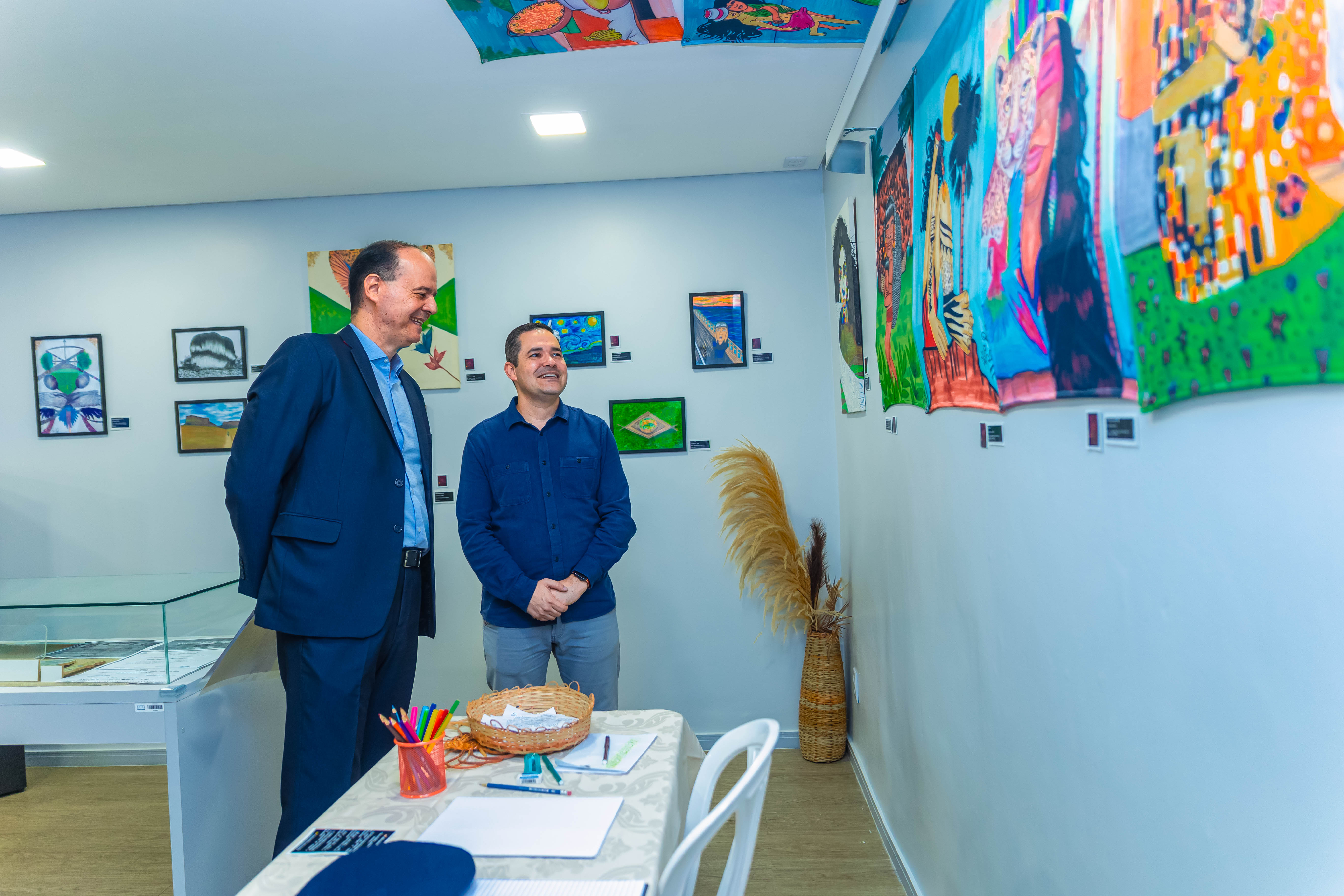 Imagem colorida do desembargador Cristóvão Suter e do Subcoordenador do CMC, Felipe Queiroz, sorriem enquanto observam pinturas coloridas penduradas em uma parede branca. Há também quadros menores emoldurados e uma mesa com materiais de desenho (lápis de cor) e um cesto. Imagem colorida do desembargador Cristóvão Suter e do Subcoordenador do CMC, Felipe Queiroz, sorriem enquanto observam pinturas coloridas penduradas em uma parede branca. Há também quadros menores emoldurados e uma mesa com materiais de desenho (lápis de cor) e um cesto.