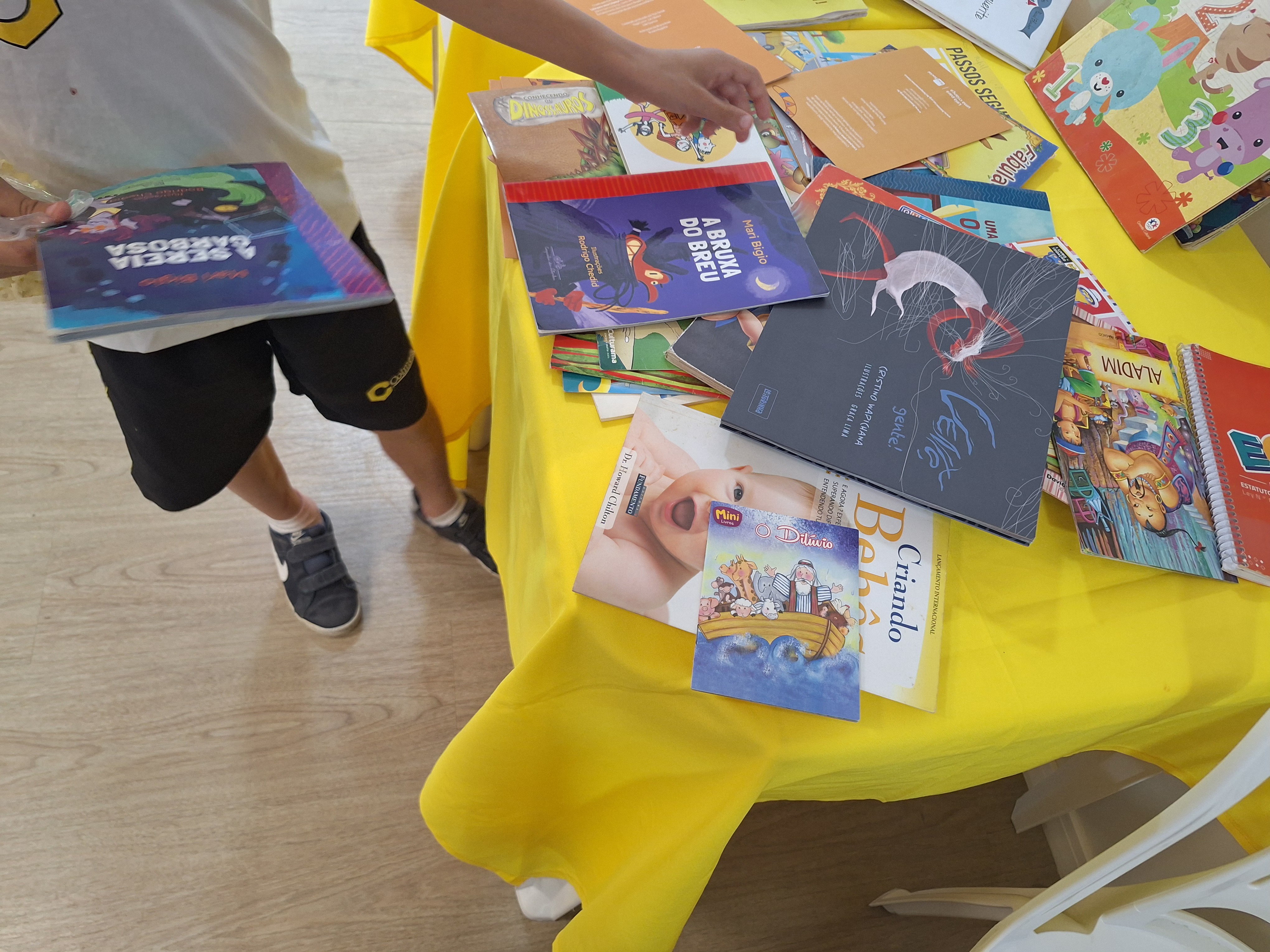 A imagem colorida mostra uma mesa coberta com toalha amarela, onde vários livros infantis estão espalhados. As capas coloridas exibem ilustrações de personagens, animais. À esquerda, uma criança em pé, vestindo camiseta branca e shorts escuros, folheia um dos livros. O ambiente é claro e transmite um clima de curiosidade e incentivo à leitura infantil.    A imagem colorida mostra uma mesa coberta com toalha amarela, onde vários livros infantis estão espalhados. As capas coloridas exibem ilustrações de personagens, animais. À esquerda, uma criança em pé, vestindo camiseta branca e shorts escuros, folheia um dos livros. O ambiente é claro e transmite um clima de curiosidade e incentivo à leitura infantil.   A imagem colorida mostra uma mesa coberta com toalha amarela, onde vários livros infantis estão espalhados. As capas coloridas exibem ilustrações de personagens, animais. À esquerda, uma criança em pé, vestindo camiseta branca e shorts escuros, folheia um dos livros. O ambiente é claro e transmite um clima de curiosidade e incentivo à leitura infantil.