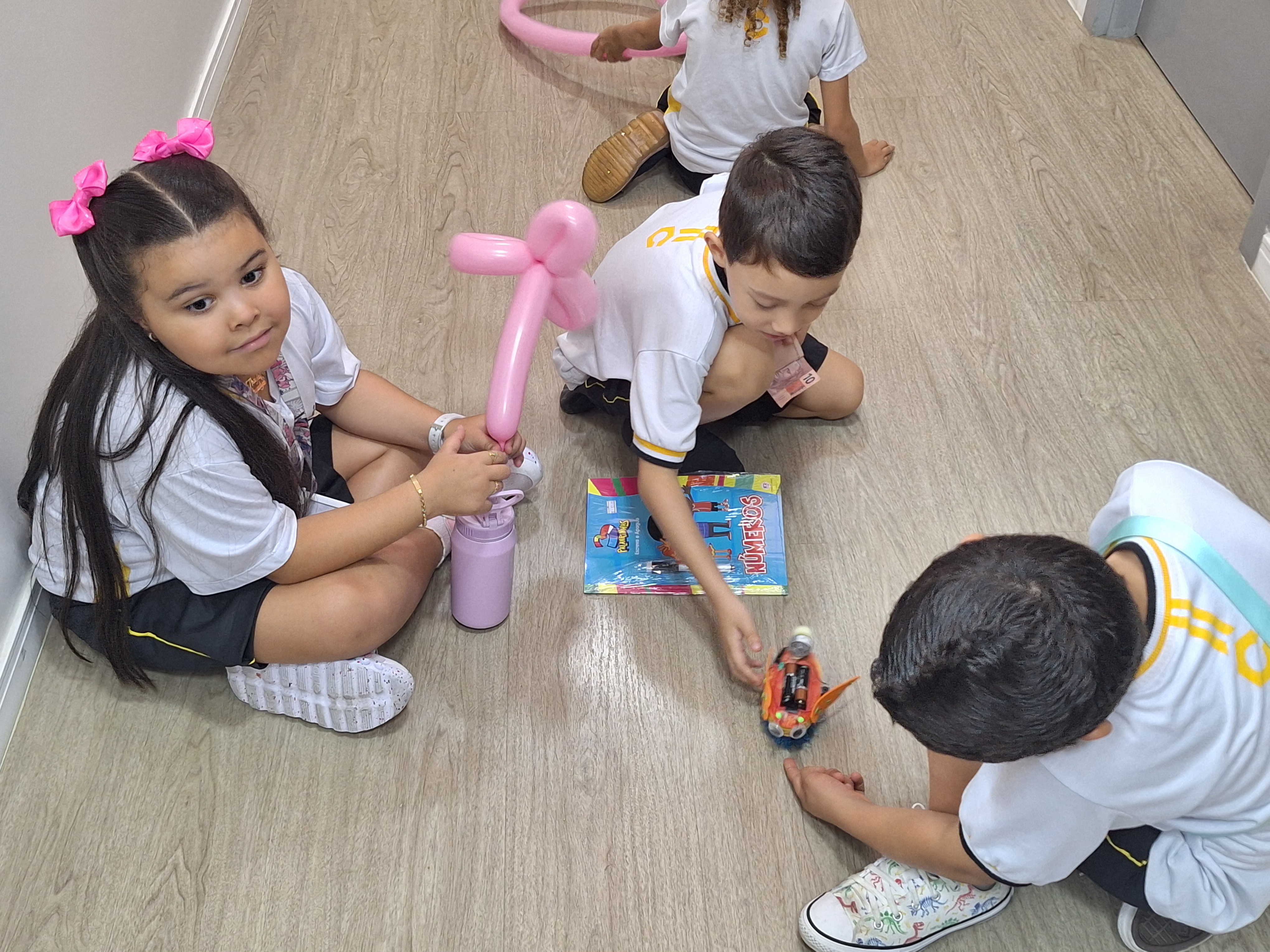 A imagem colorida mostra quatro crianças sentadas no chão de um corredor de piso claro, brincando e interagindo. À esquerda, uma menina com longos cabelos escuros, laços cor-de-rosa na cabeça e uniforme escolar segura um balão modelado em forma de flor e uma garrafinha rosa. Ao lado, um menino observa um carrinho de brinquedo enquanto segura um livro colorido. Outras duas crianças também participam da brincadeira, concentradas no carrinho. O ambiente é iluminado e transmite um clima de descontração e alegria infantil.   A imagem colorida mostra quatro crianças sentadas no chão de um corredor de piso claro, brincando e interagindo. À esquerda, uma menina com longos cabelos escuros, laços cor-de-rosa na cabeça e uniforme escolar segura um balão modelado em forma de flor e uma garrafinha rosa. Ao lado, um menino observa um carrinho de brinquedo enquanto segura um livro colorido. Outras duas crianças também participam da brincadeira, concentradas no carrinho. O ambiente é iluminado e transmite um clima de descontração e alegria infantil.